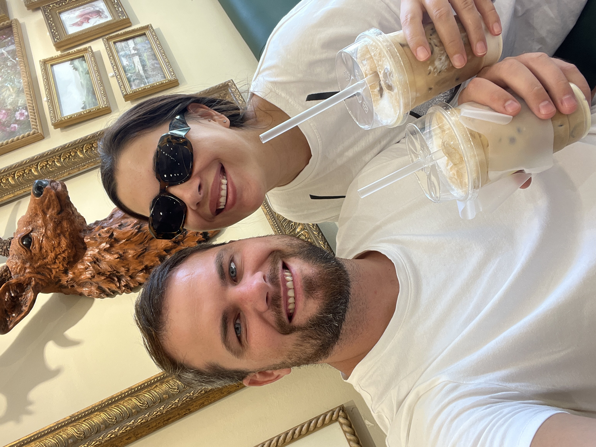A man and a woman are posing for a selfie while holding iced coffee drinks. The man, in the foreground on the left, has fair skin, blue eyes, and a beard. He is wearing a white t-shirt. The woman, on the right, has dark hair, fair skin, and is wearing large, dark sunglasses and a white t-shirt. She is smiling. In the background, there are framed pictures on the wall and a taxidermied deer head. The woman is holding one iced coffee, and the man is holding another. The scene appears to be indoors, possibly in a cafe or restaurant. The image does not provide specific details to identify the location within Nha Trang Ward, Vietnam.