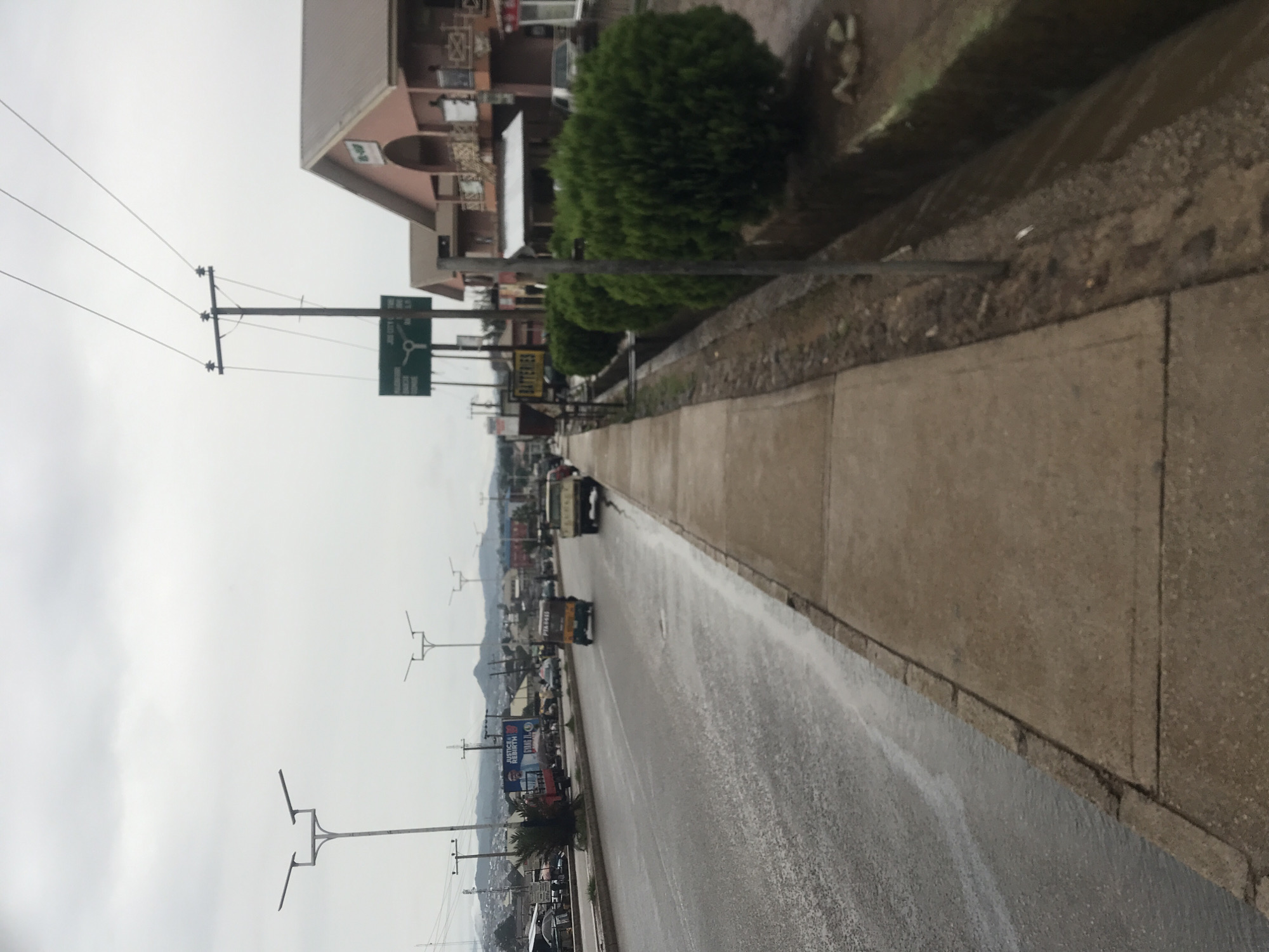 The image displays a street scene from an urban or semi-urban area, likely in Jos, Nigeria, viewed from the side of the road looking into the distance. The sky is overcast and gray, indicating a cloudy day, and the asphalt road is visibly wet, suggesting recent rainfall.
On the right side of the frame, a concrete sidewalk runs alongside the wet road, separated by a curb and a narrow strip of dirt and low-lying green bushes. Beyond this, a row of multi-story buildings, some with balconies, line the street. Utility poles with multiple overhead wires are prominent along the left side of the road.
Various vehicles, including cars and what appear to be motorcycles or three-wheeled vehicles, are present on the road. In the mid-ground, a green directional sign mounted on a utility pole features a white roundabout symbol. Text on this sign clearly indicates "JOS CITY," "PLATEAU STADIUM," "FEDERAL SECRETARIAT," and "BASHIRU" (partially visible). Further down the street, a yellow sign advertising "BATTERIES" is visible. Distant billboards and commercial signs can be seen, one of which is blue and prominently displays "JUSTICE & REBIRTH" and "BUILD THE..." A yellow vehicle in the distance also shows the alphanumeric text "PTN 9465". The scene captures everyday activity along a main thoroughfare.