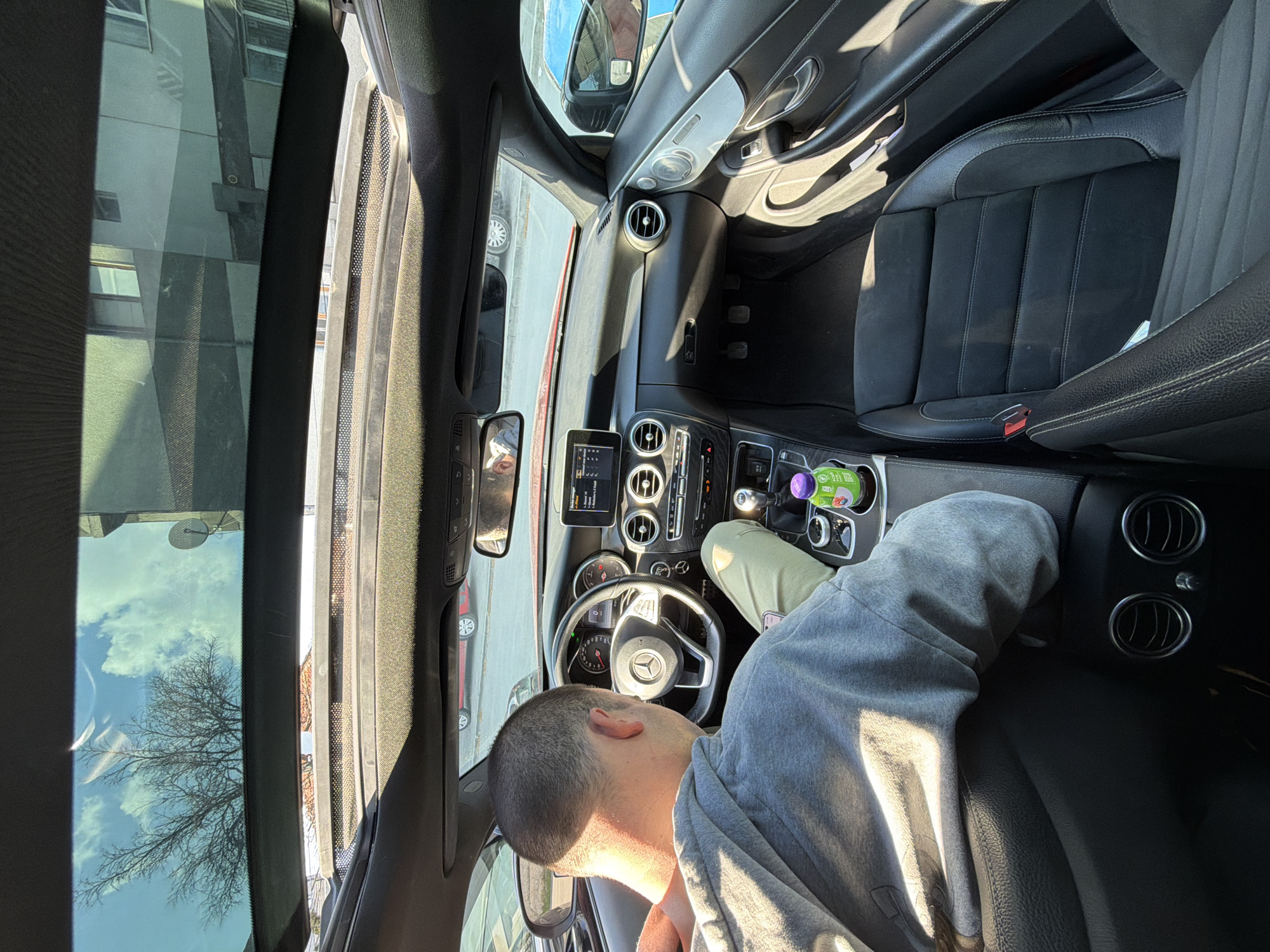 The image, rotated 90 degrees clockwise, captures the interior of a car, focusing on the driver's area and dashboard. A person with short, dark hair is seated in the driver's seat, wearing a grey hooded sweatshirt and light-colored pants. Their head is seen from the back, and their left leg is casually propped up against the dashboard near the center console.
The car's interior features black leather seats with ribbed sections, a Mercedes-Benz steering wheel, a modern dashboard with multiple circular air vents, and an infotainment screen displaying unreadable digital information. A gear shifter is visible, and a purple-capped bottle with the text "PURE" and other unreadable words sits in a cupholder. The car has a panoramic sunroof, reflecting a clear blue sky with white clouds and the silhouette of a bare tree, suggesting late autumn, winter, or early spring.
Outside the car, visible through the windows and reflections, is an urban setting in Varna, Bulgaria. Buildings with multiple windows are reflected, along with parts of other parked vehicles, including a red car. Bright sunlight casts strong shadows within the car, indicating a sunny daytime. The person appears to be resting or waiting in a relaxed posture inside the parked car. No specific emotions are discernible from their partially visible face.
