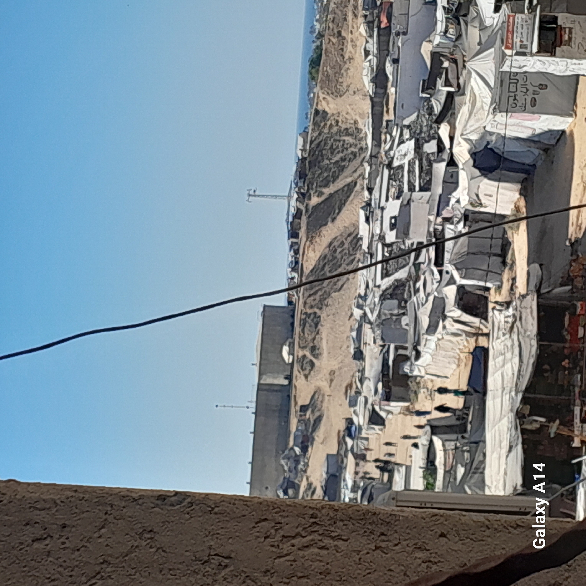 The image shows a densely populated refugee camp, possibly in Khan Yunis, Palestinian Territories. Numerous tents and makeshift shelters are visible, packed closely together. People are seen walking amongst the structures, suggesting an active community. In the background, a large, imposing concrete structure sits atop a hill, overlooking the camp and the sea. The overall scene depicts a challenging living environment, common in refugee settlements.