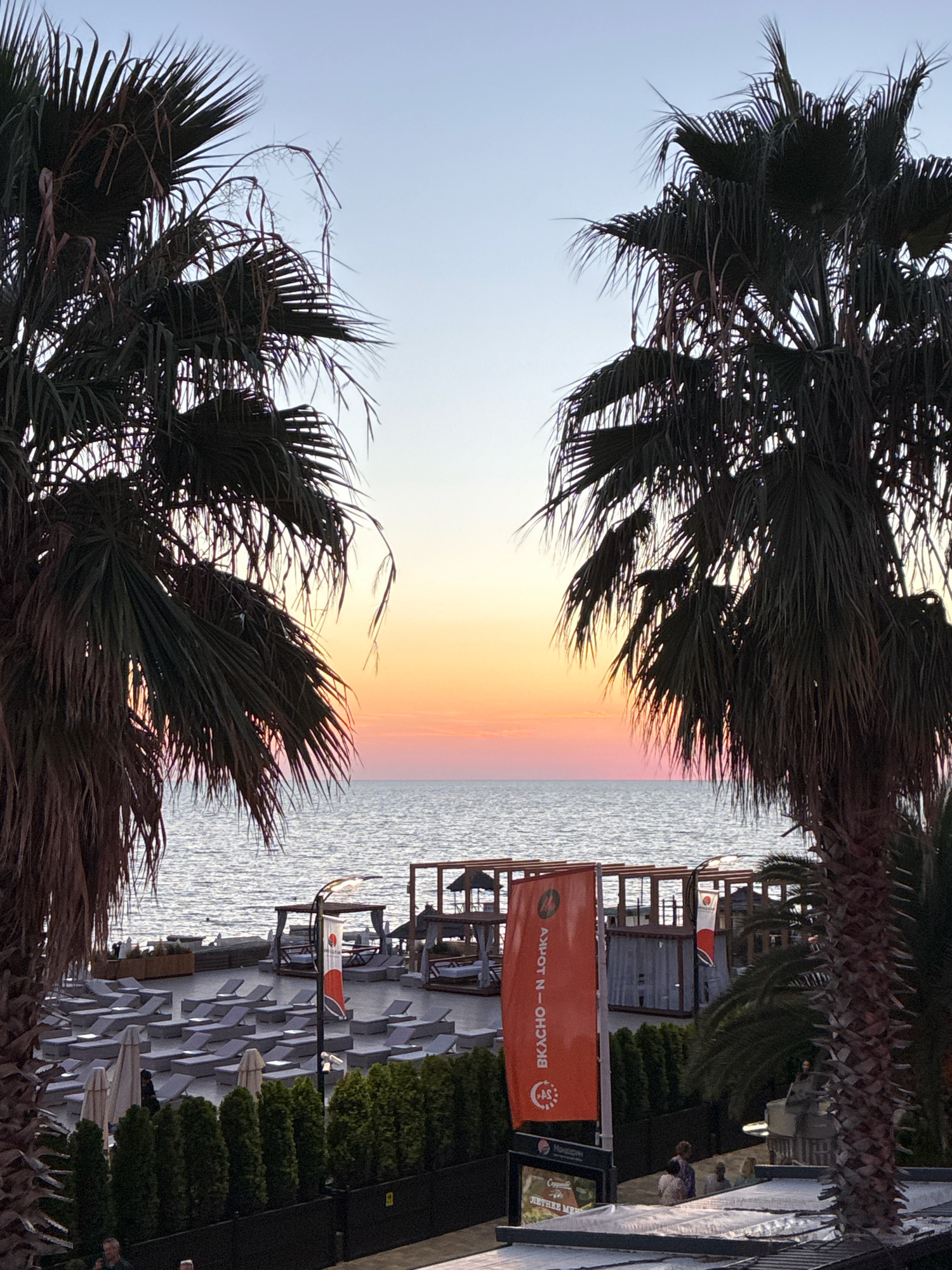 This image depicts a serene sunset scene at a beach resort in Sochi, Russia. The sky transitions from a pale blue at the top to a warm gradient of orange and pink near the horizon, reflecting softly on the calm sea. In the foreground, two large palm trees frame the view, their dark fronds silhouetted against the colorful sky.
The beach area is meticulously arranged with numerous empty lounge chairs and umbrellas, suggesting a well-maintained resort. Several cabanas and structures are visible further down the beach. An orange flag with Cyrillic text, likely a brand or promotional banner, stands prominently near the center, next to a lamppost. A few people are scattered throughout the resort area, some seated and others walking, appearing relaxed and enjoying the evening ambiance. The overall impression is one of tranquility and leisure.