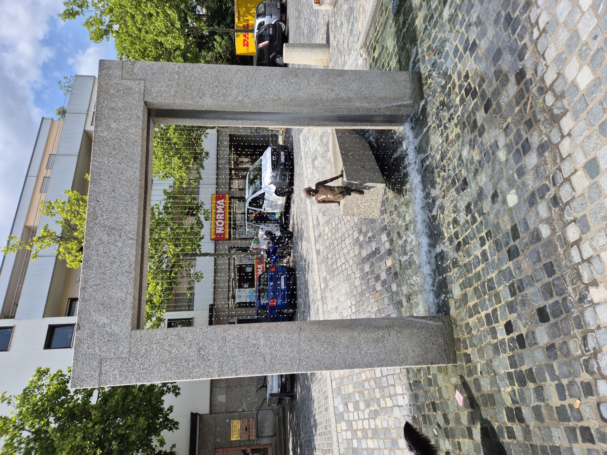 The image depicts a public square in Regensburg, Germany, featuring a stone archway with a water feature. A small bronze statue of a child is seated on the edge of the water feature, with water splashing around it. The ground is paved with cobblestones. In the background, a building with a "NORMA" grocery store is visible, along with parked vehicles including a blue Volkswagen and a white van. Trees with green foliage line the square, and the sky is partly cloudy. A black dog is partially visible in the lower left corner.