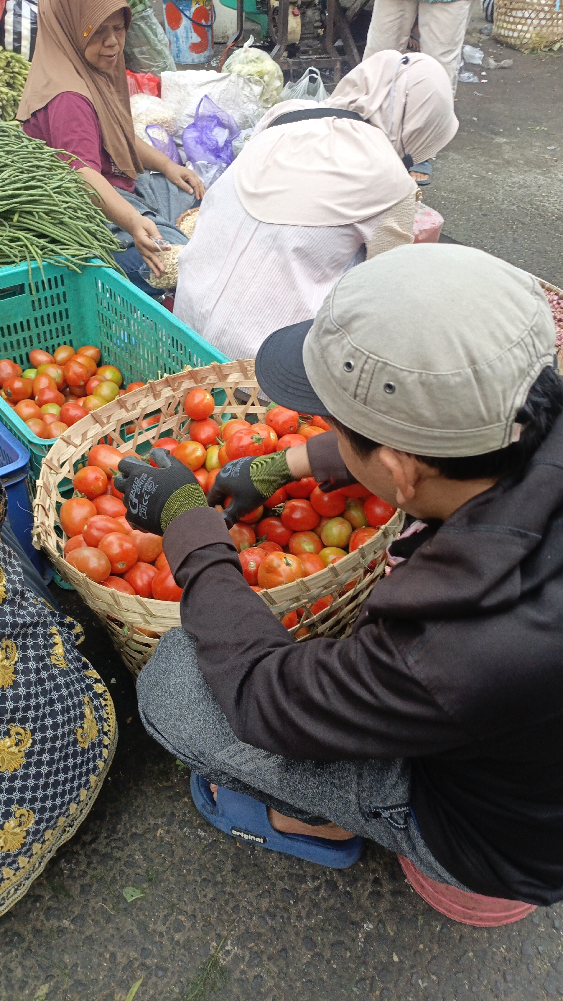 A market stall in Salatiga, Indonesia, displays an abundance of fresh produce. In the foreground, a person wearing a cap and black gloves is sorting through a basket overflowing with ripe red tomatoes. To the left, a green plastic crate holds a pile of long green beans, and behind it, another person wearing a headscarf is attending to what appears to be a basket of dried goods. Further back, another individual in a pale headscarf and striped top is also engaged in market activities. The ground is paved and appears damp, suggesting recent rain or cleaning. The scene captures a typical moment of commerce at an Indonesian market.