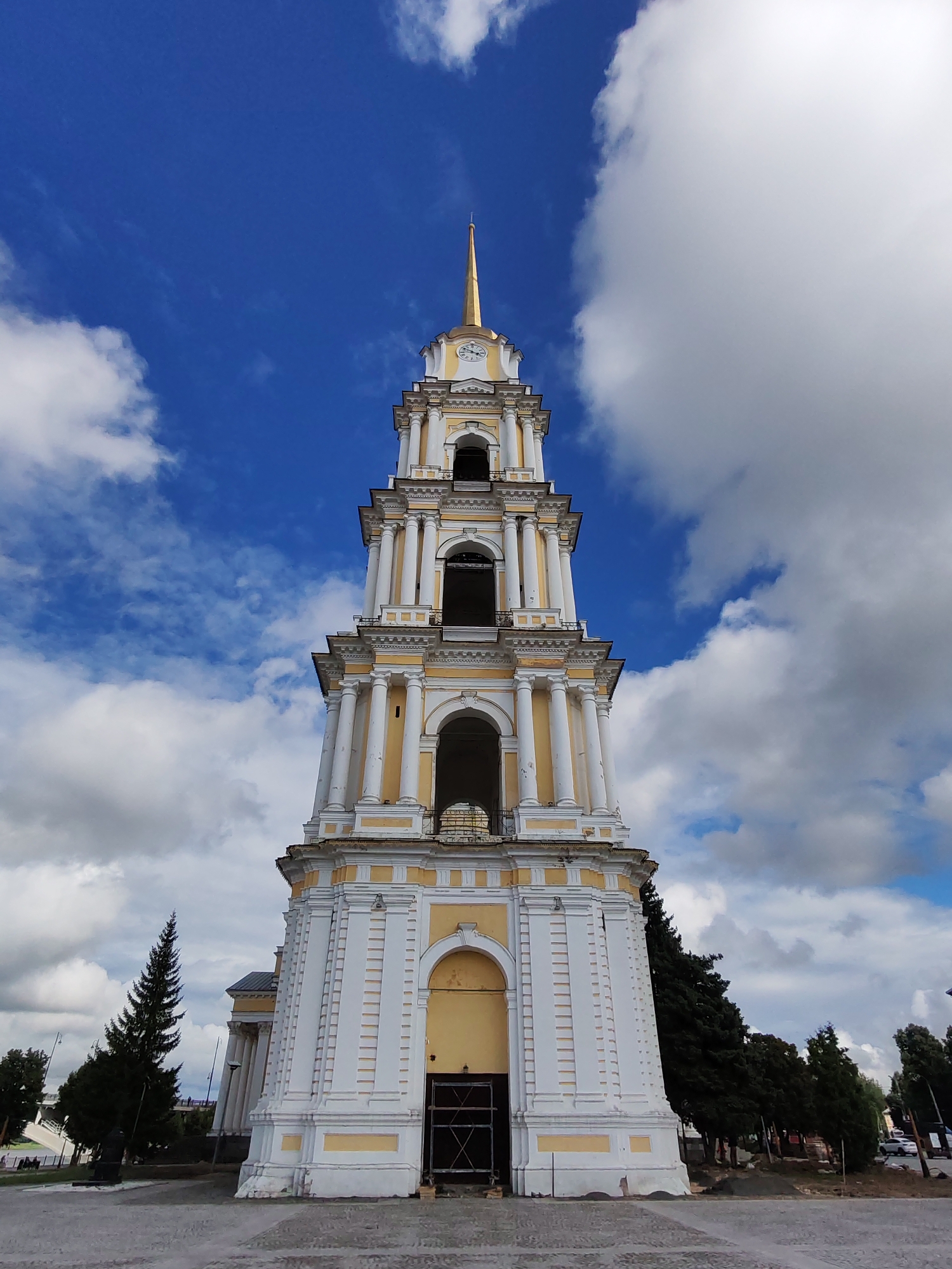 A low-angle, full shot captures the imposing white and yellow bell tower of the Church of Saints Boris and Gleb in the city of Bol'shie Mkhi, Russia, against a dynamic blue sky filled with scattered white clouds. The bell tower, adorned with classical architectural elements such as columns and arched openings, culminates in a gold spire topped with a cross. A clock face is visible on one side of the tower. The base of the tower features large dark doors. To the left of the tower, a tall, dark green coniferous tree stands prominently. The ground in the foreground is paved with cobblestones. Further back, to the left, a section of another building with a white facade and columns is visible, as well as a bridge structure and people walking. To the right of the bell tower, a line of dark green trees forms a backdrop. The overall scene conveys a sense of historical architecture under a partly cloudy sky.