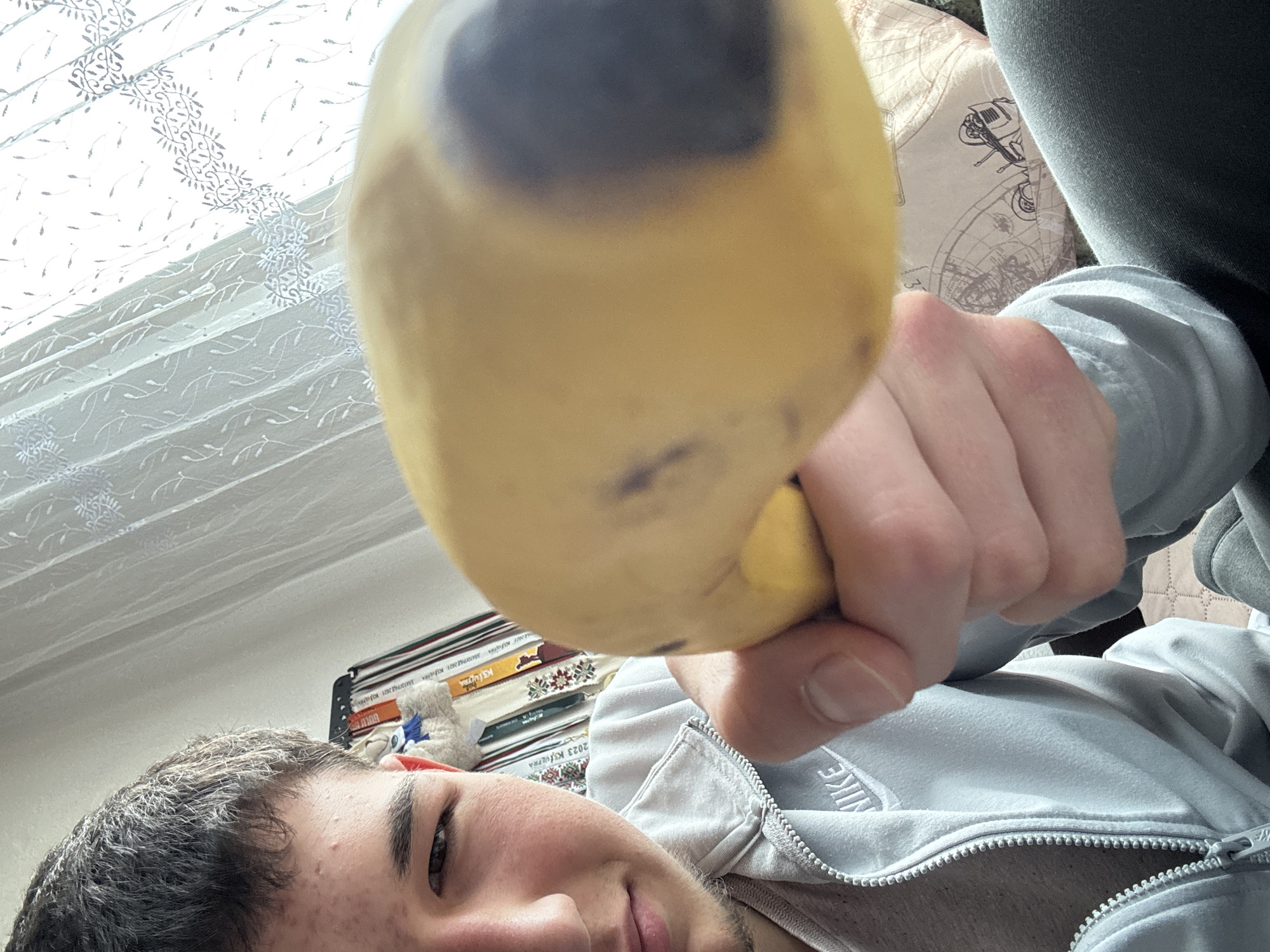 A young man is holding a bruised yellow mango in front of his face. He is wearing a light gray Nike zip-up hoodie. He is inside, with a stack of books visible behind him, one titled "2023 KUFIRA". A patterned curtain is visible near a window. The location is Zlatograd, Bulgaria.