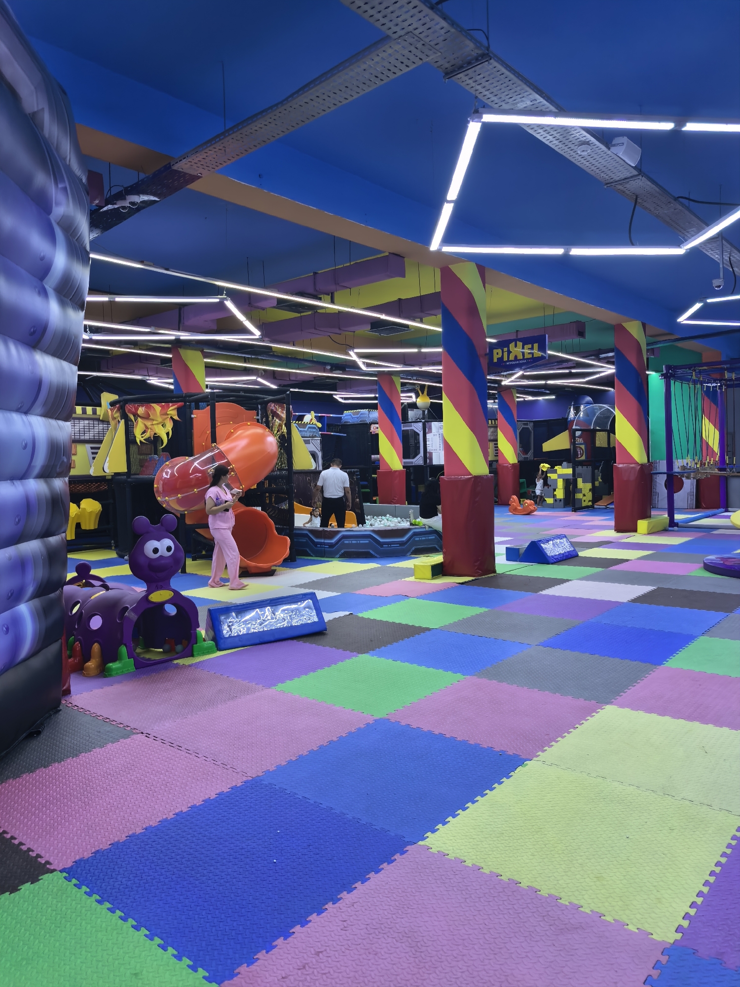 The image depicts the interior of a brightly colored indoor playground, likely located in Fergana City, Uzbekistan. The floor is covered with interlocking foam mats in a checkerboard pattern of various colors, including blue, purple, pink, green, and yellow. In the foreground on the left, a large, inflated purple structure with a textured surface is visible. To its right are two purple play structures shaped like cartoon insects with wheels and openings. A young child in pink clothing stands near an orange slide and play structure, holding a tablet. In the background, a man in a white shirt and dark pants is observed. Above, the ceiling is painted blue and adorned with colorful beams and modern geometric light fixtures. Pillars with red, yellow, and blue spiral patterns support the ceiling. A sign reading "PIXEL" with text below in a Cyrillic script is visible on one of the pillars. Further in the background, elements of a large play structure with slides, climbing nets, and yellow and black modules are present.
