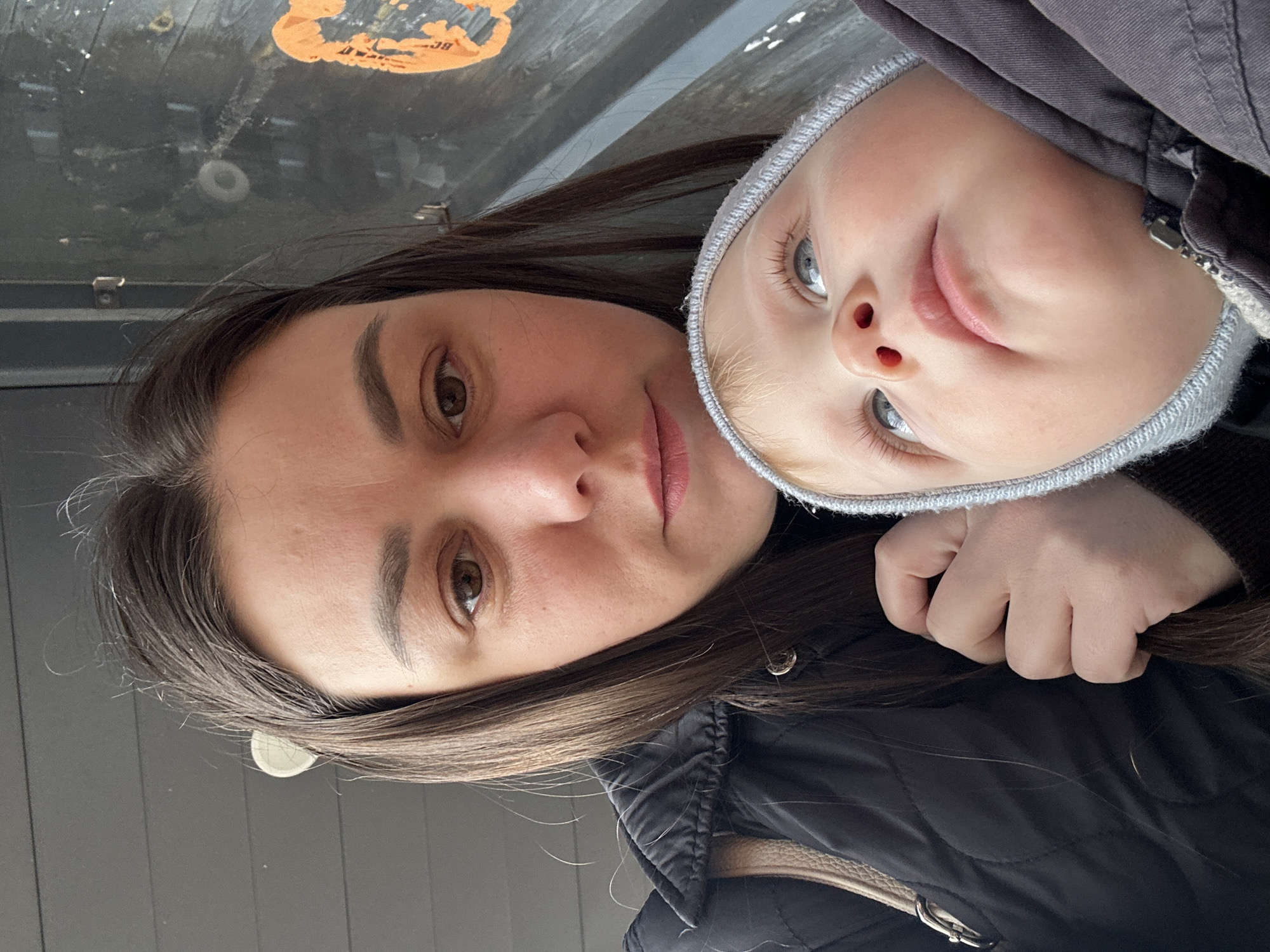 A woman and a young child are posing for a selfie. The woman has long, dark brown hair and brown eyes. She is wearing a dark quilted jacket. The child has blonde hair, blue eyes, and is wearing a grey hat and a dark jacket. The child is holding the woman's hair with their hand. The background appears to be a building or structure with a gray exterior and an older sticker. It is likely taken outside in Chaikovsky, Russia.