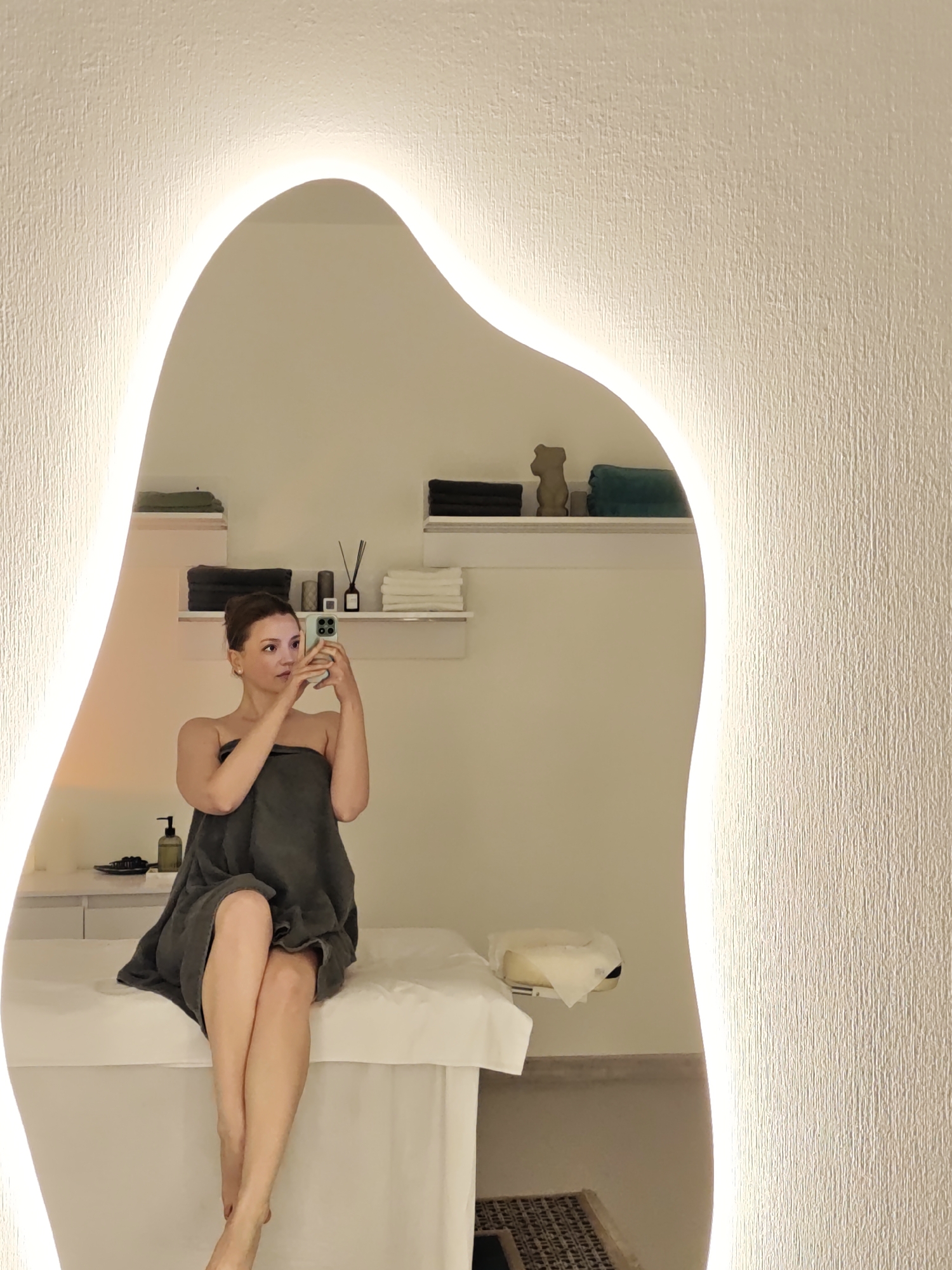 A young woman, wrapped in a dark gray towel, sits on the edge of a white treatment table. She is holding a smartphone and taking a selfie. The scene is a spa or treatment room. Behind her, a white counter holds toiletries. Shelves above hold rolled towels, decorative objects, and aroma diffuser sticks. The reflection is captured in an irregularly shaped mirror with an illuminated border. The background shows the textured wall of the spa, and a glimpse of the floor. The scene likely takes place in Vinnytsia, Ukraine.