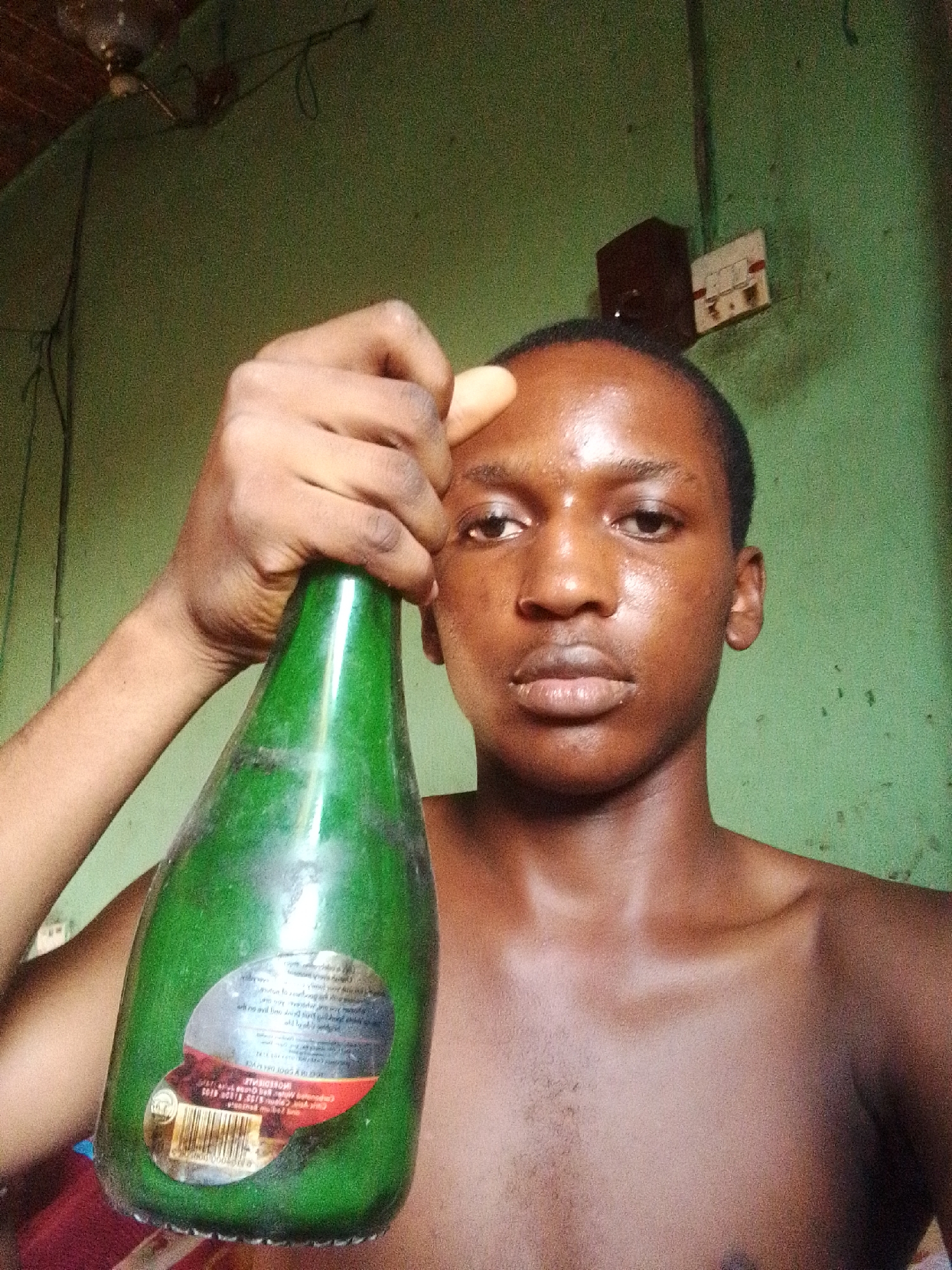 A medium shot shows a young Black person holding a green glass bottle with a label in the city of Nkpor, Nigeria. The person is shirtless, with their face centered in the frame. They have dark skin, short black hair, and are looking directly at the camera. Their right hand is holding the neck of the bottle, which is tilted downwards and in front of their chest. The background consists of a textured green wall with an electrical outlet and a light fixture visible near the top of the frame. The lighting is dim, casting a glow on the person's skin. The overall impression is of a casual, personal snapshot.