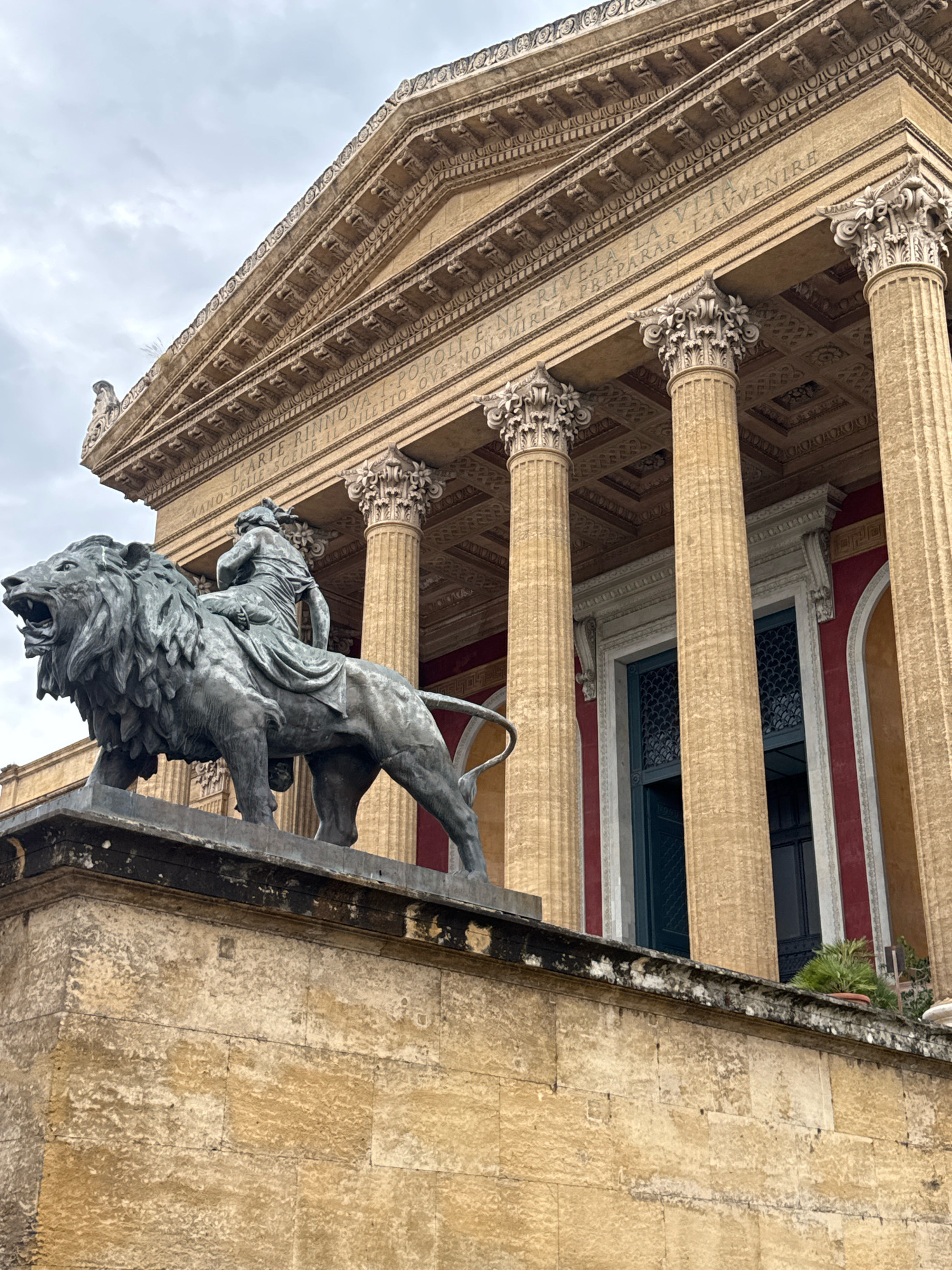 The image shows the Teatro Massimo in Palermo, Italy, a grand opera house with neoclassical architecture. In the foreground, a weathered stone pedestal supports a bronze statue of a lion with a person riding on its back. The lion is depicted with its mouth open, as if roaring, and its mane is detailed and flowing. The rider appears to be a draped female figure. Behind the statue, the facade of the Teatro Massimo is visible, characterized by large, fluted Corinthian columns supporting a pediment. Inscriptions are visible on the frieze above the columns. The building has a red section behind the columns, and tall, dark-paned windows. The sky is overcast, suggesting a cloudy day. The overall impression is one of historical grandeur and artistic monumentality.