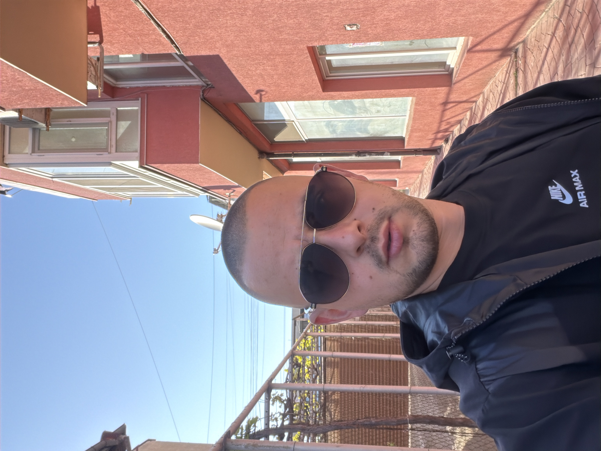 A medium shot depicts a light-skinned male wearing sunglasses and a black Nike Air Max jacket. He is positioned outside, with a weathered metal railing visible below. Behind the railing, greenery is observed. The backdrop includes a red-brick building, clear blue sky, and a satellite dish. The city and specific location are unknown.