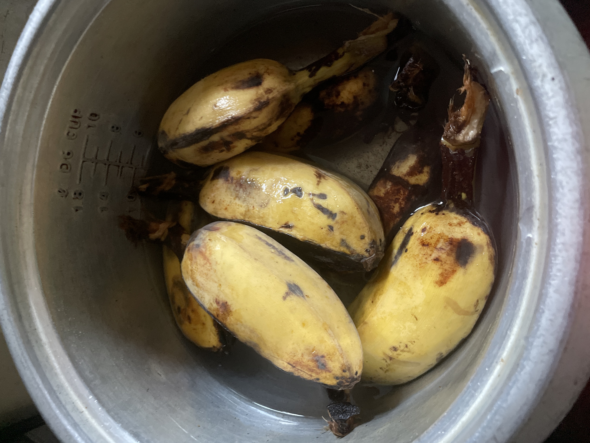 The image shows several ripe plantains submerged in water inside a metal cooking pot. The plantains are yellow with some brown spots and darken toward the stem. The pot appears to be the insert from a rice cooker, judging by the visible measurement markings on the inner surface. The picture was likely taken indoors. There is no information in the image to confirm a location within Davao City, Philippines.