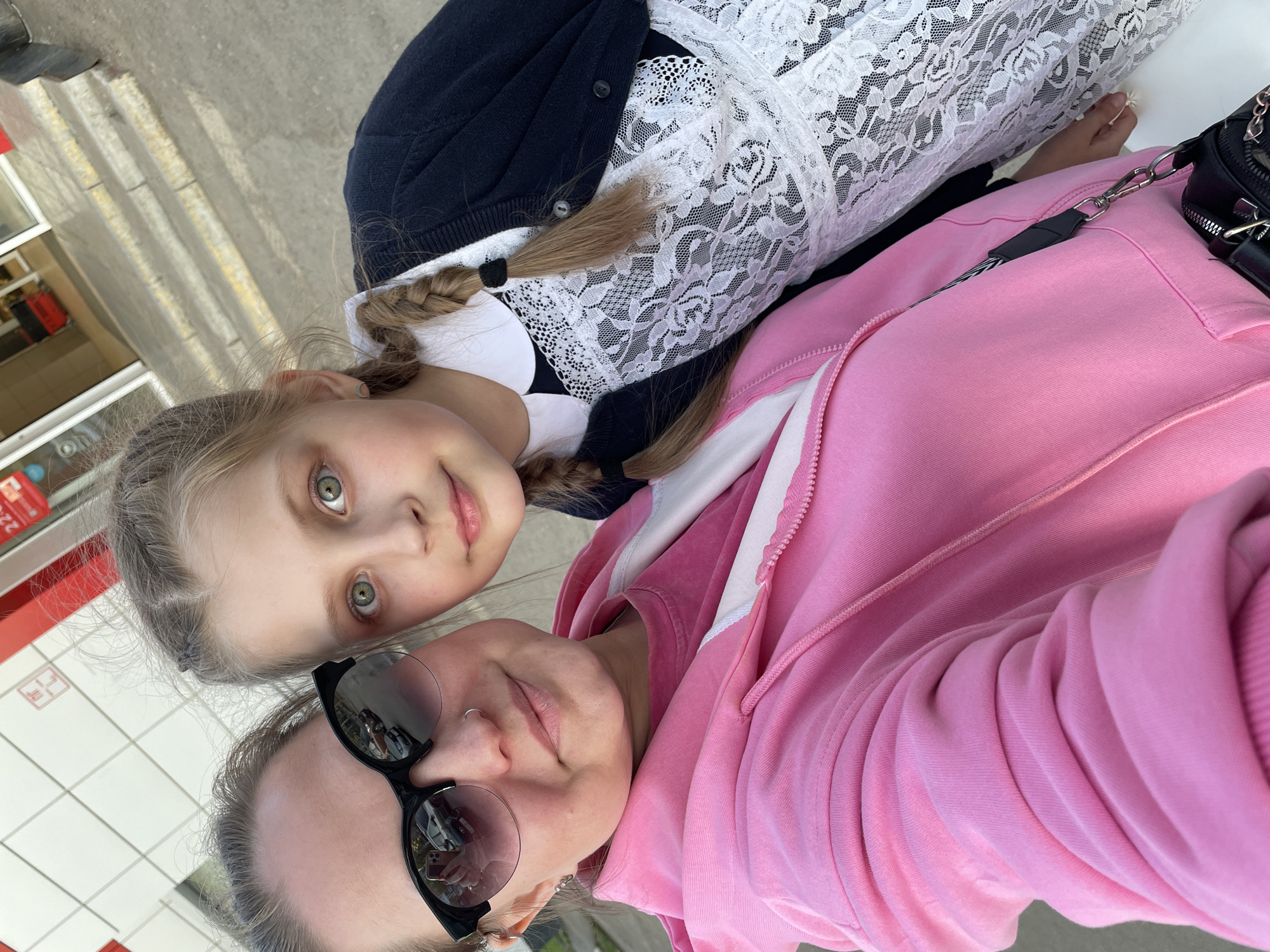A selfie shows a young girl with blonde braids and green eyes looking at the camera. She is wearing a dark blue cardigan and a white lace collar. Beside her is an adult woman with dark hair and sunglasses wearing a pink hoodie. They are standing outside, possibly in front of a building with white and red tiled walls. Reflections of cars are visible in the woman's sunglasses. The location is likely Izhevsk, Russia, based on the presence of tiled walls that are common there.