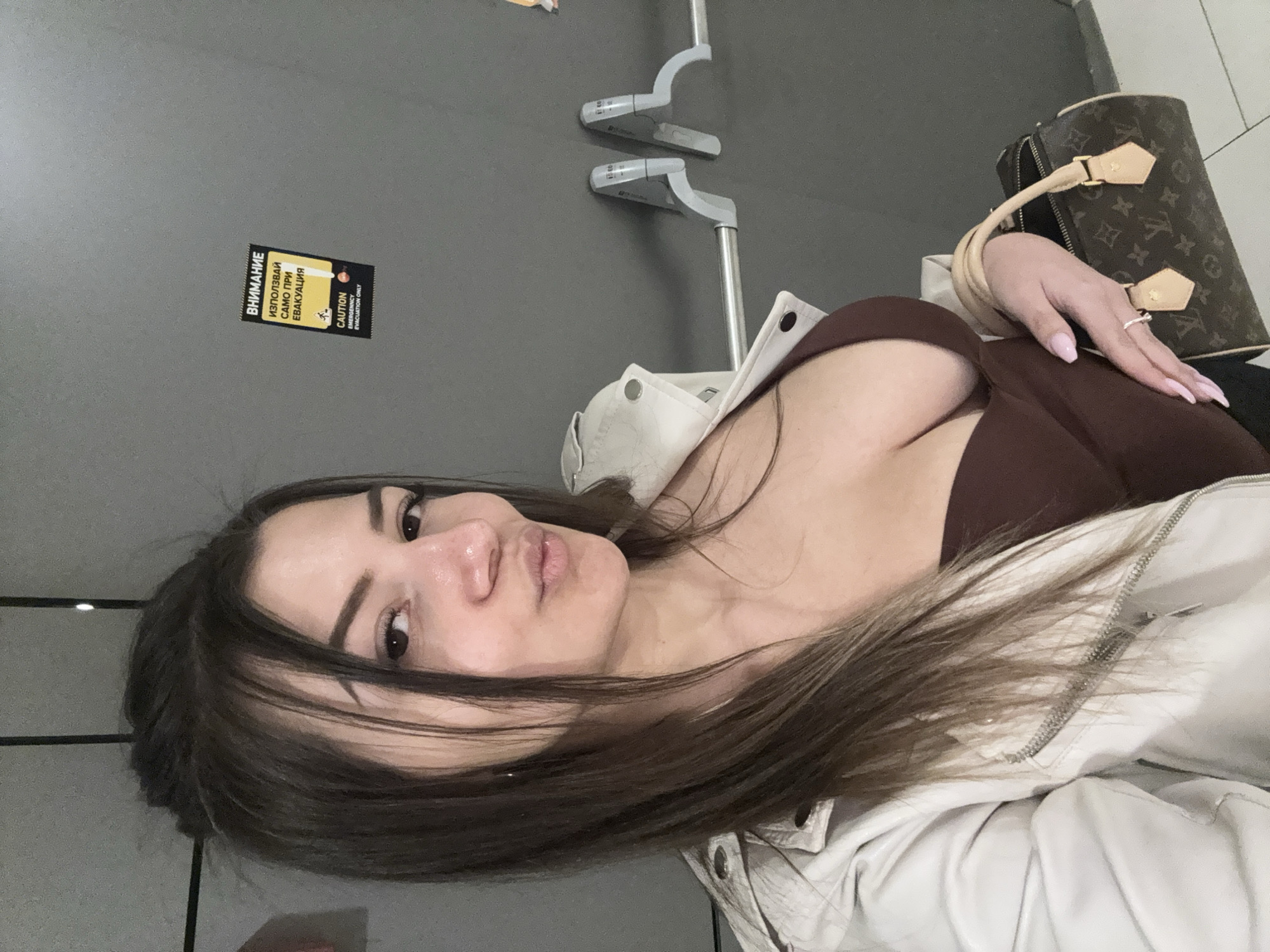 A young woman with long brown hair and fair skin is depicted in a selfie, taken at an angle. She is wearing a brown top and a cream-colored jacket. Her manicured hand with light pink nails and a ring rests near a patterned handbag. The background includes a gray wall with a sign in Russian or Bulgarian that includes the word "эвакуация" (evacuation). Above are gray metal rails. The location is identified as Sofia, Bulgaria.