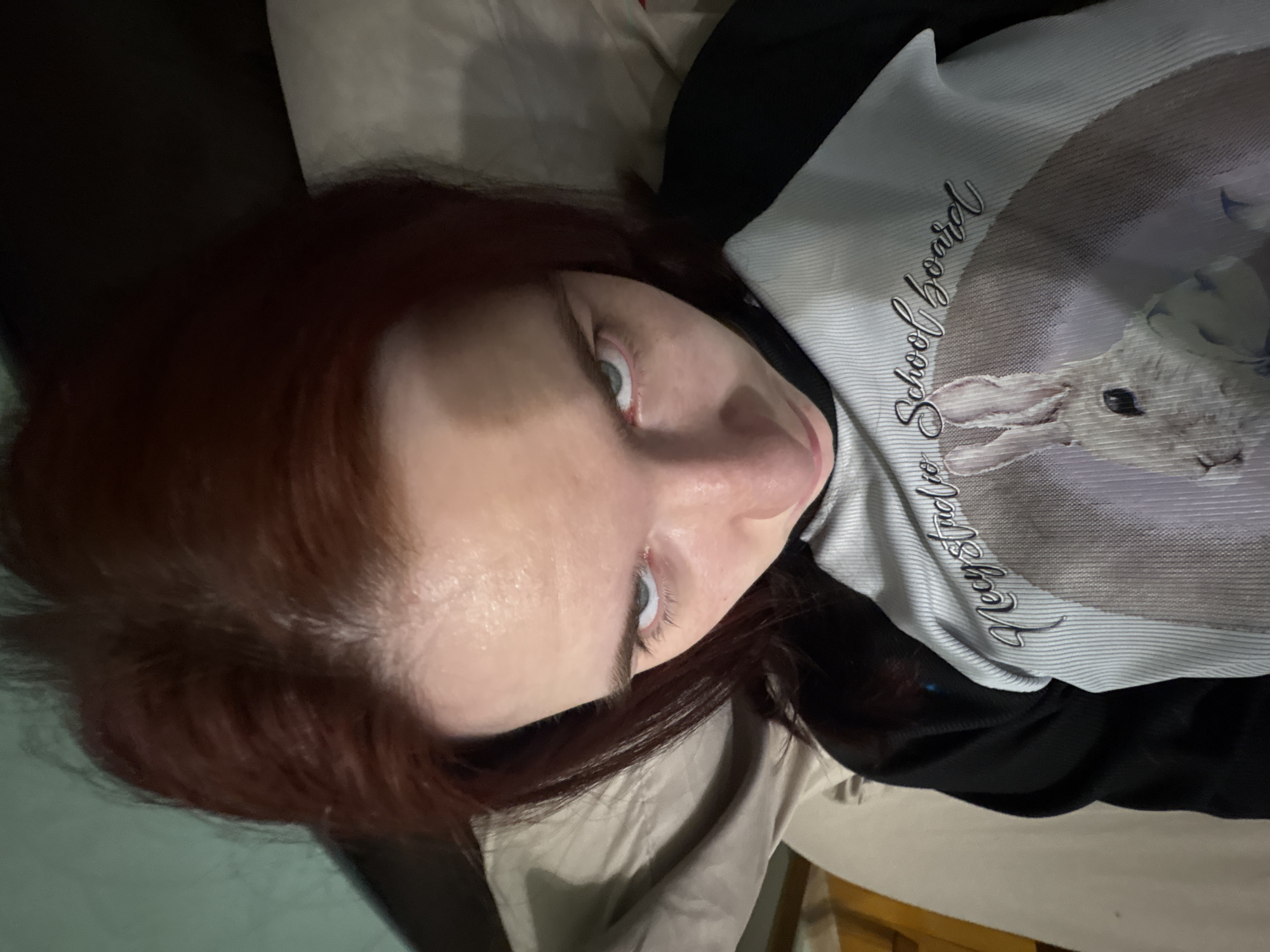 A young woman with reddish-brown hair lies in bed, looking directly at the camera. Her eyes are wide open and her eyebrows are furrowed, giving her an intense expression. She is wearing a black and white raglan t-shirt with an image of a rabbit and text that reads "Negy Studio School Board" embroidered on the front. The background is slightly out of focus, showing a pillow and the side of a bed. The lighting is dim and appears to be coming from an overhead source. The woman is positioned in the city of Sinyavino, Russia.