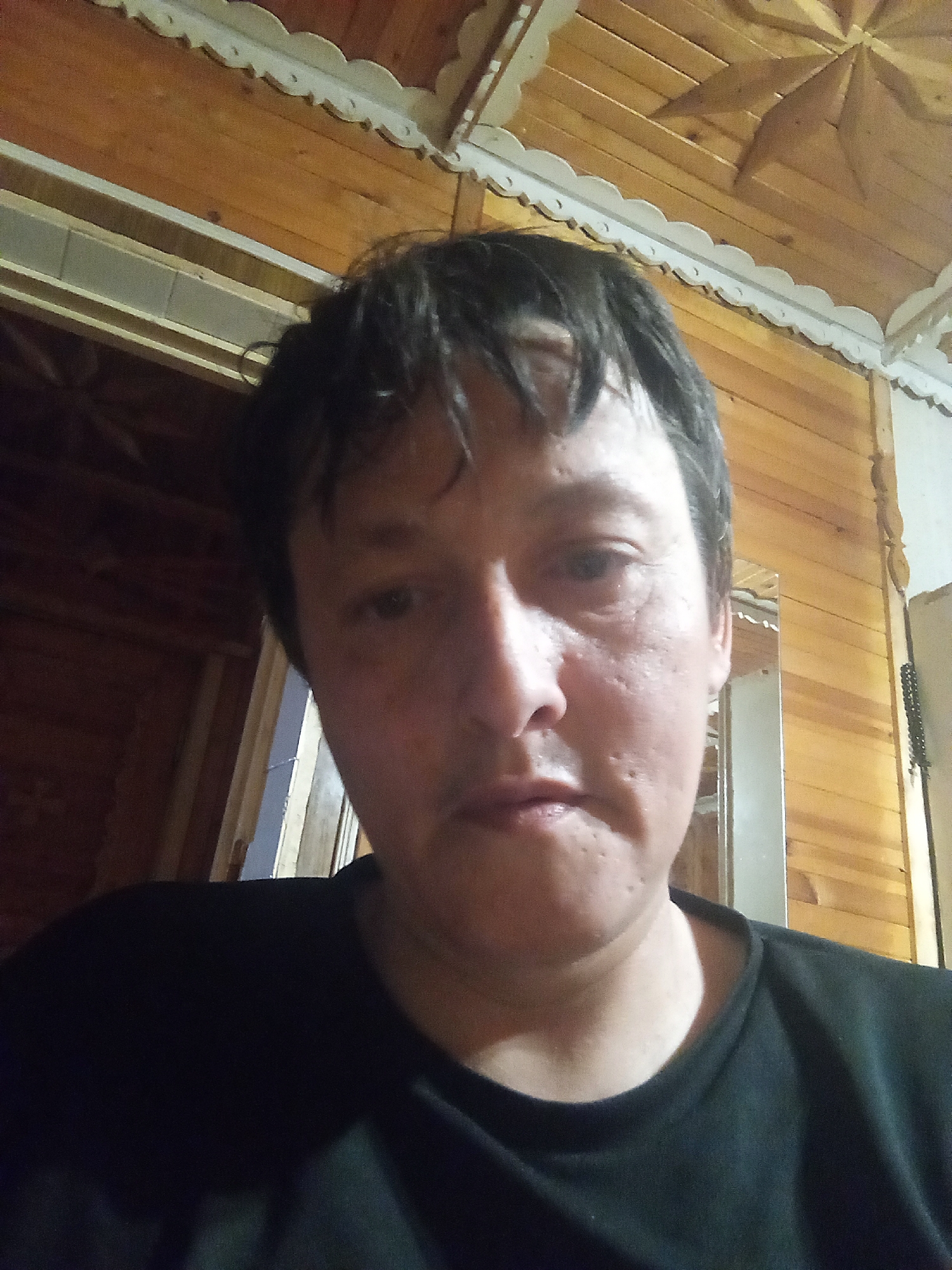 A person with short, dark hair and fair skin is captured in a close-up, selfie-style photograph. They are wearing a black t-shirt. The background suggests an indoor setting with wooden paneling on the walls and ceiling, which features decorative trim and a star-shaped design on the ceiling. The lighting appears to be dim and warm. The image lacks specific context that would allow for the identification of the exact location within Vsevolozhsk, Russia.
