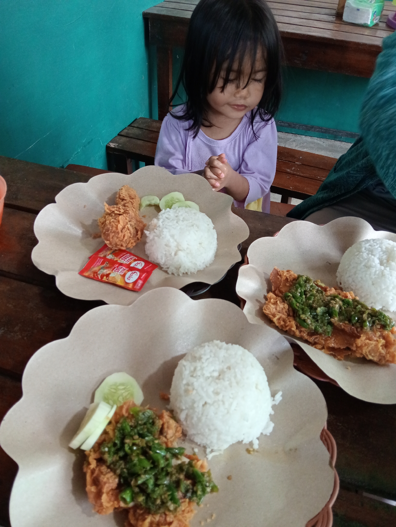 In Campago, Indonesia, a young girl with black hair is seated at a wooden table, her hands clasped together. She is wearing a light purple shirt. In front of her are two plates of food, each containing a portion of fried chicken, white rice, and cucumber slices. One plate also has a small packet of red chili sauce. A second person, partially visible on the right side of the frame, is wearing a dark green garment. The background consists of a turquoise wall and a wooden bench. The setting appears to be an outdoor or casual dining area.