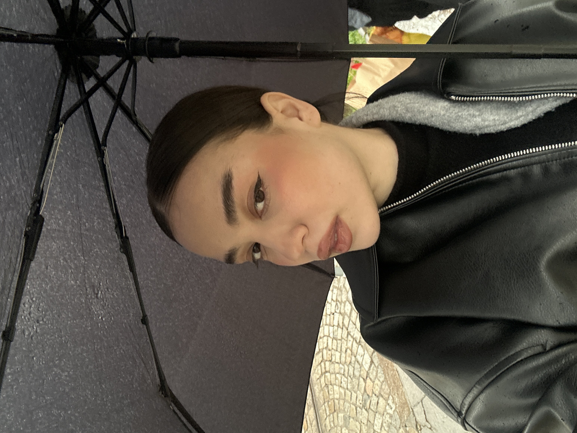 A young woman is photographed at close range from a low angle, under a black umbrella. She has dark hair pulled back, light skin, and wears makeup including blush and eyeliner. She is wearing a black leather jacket with a grey hoodie underneath. The background shows a cobblestone street. Based on the cobblestone street and architectural style, the location is likely the city of Sofia, Bulgaria.