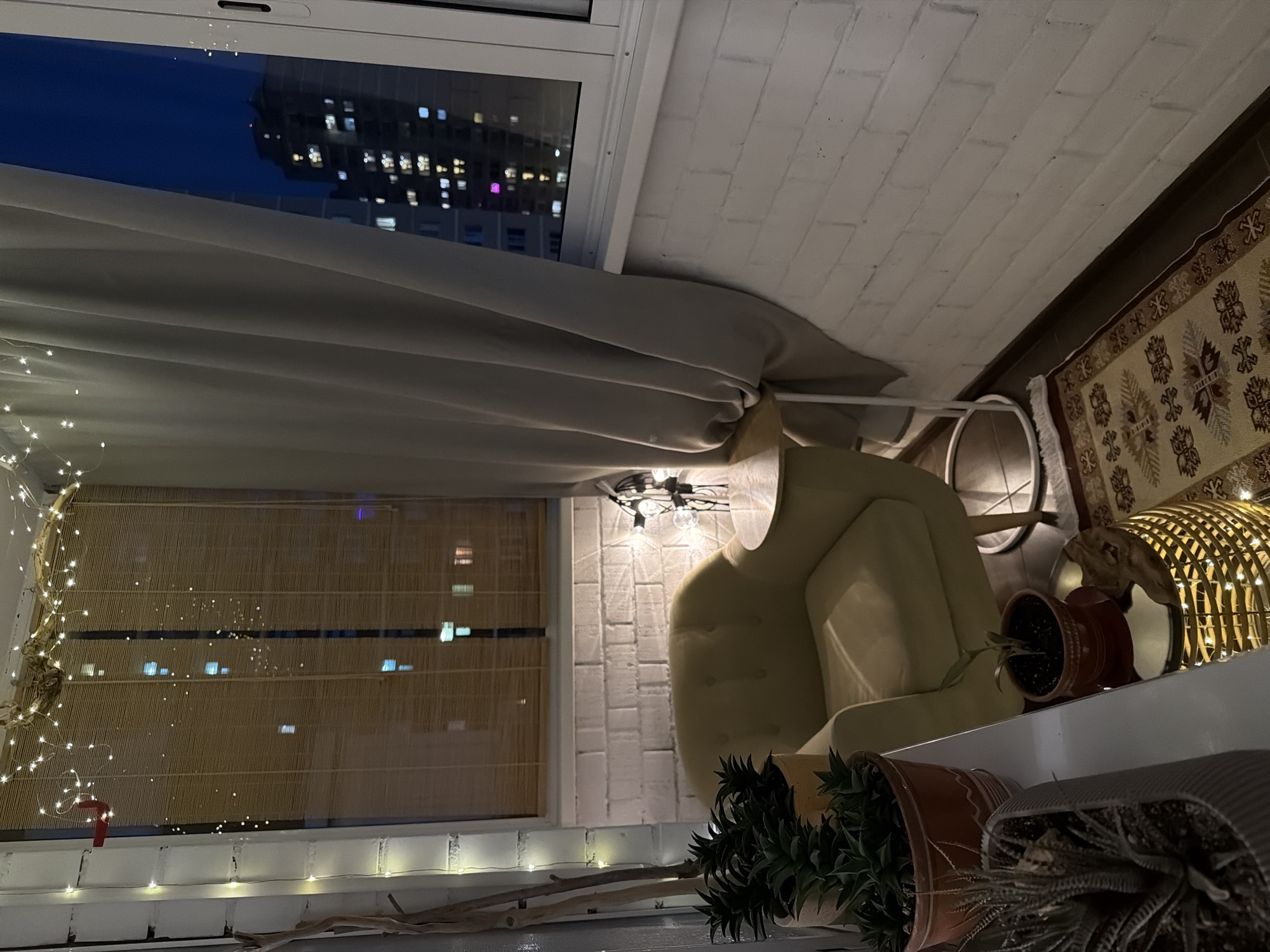 Here is a detailed description of the image:
**Location:** Unknown city, Unknown location. The scene appears to be an enclosed balcony or sunroom.
**Environment:** The space is illuminated by a combination of artificial light (likely from a light fixture and string lights) and ambient city light visible through windows. The walls are white brick, suggesting an interior space.
**Objects:**
* **Furniture:** A light-colored armchair is a prominent feature.
* **Decor:** There are several potted plants, some with greenery and others with what appears to be succulents. A woven carpet adds texture. String lights adorn the balcony. There's a decorative lamp with a cage.
* **Window Coverings:** There are light grey curtains and woven blinds.
* **Window View:** A window reveals a dark sky with a view of a distant cityscape at night.
**Subjects:** There are no people immediately visible.
**Actions/Interactions:** There is no overt action occurring.
**Essential Details:** The overall impression is of a cozy, enclosed balcony space used as a relaxation area. The combination of plants, soft lighting, and soft furnishings contributes to this atmosphere.