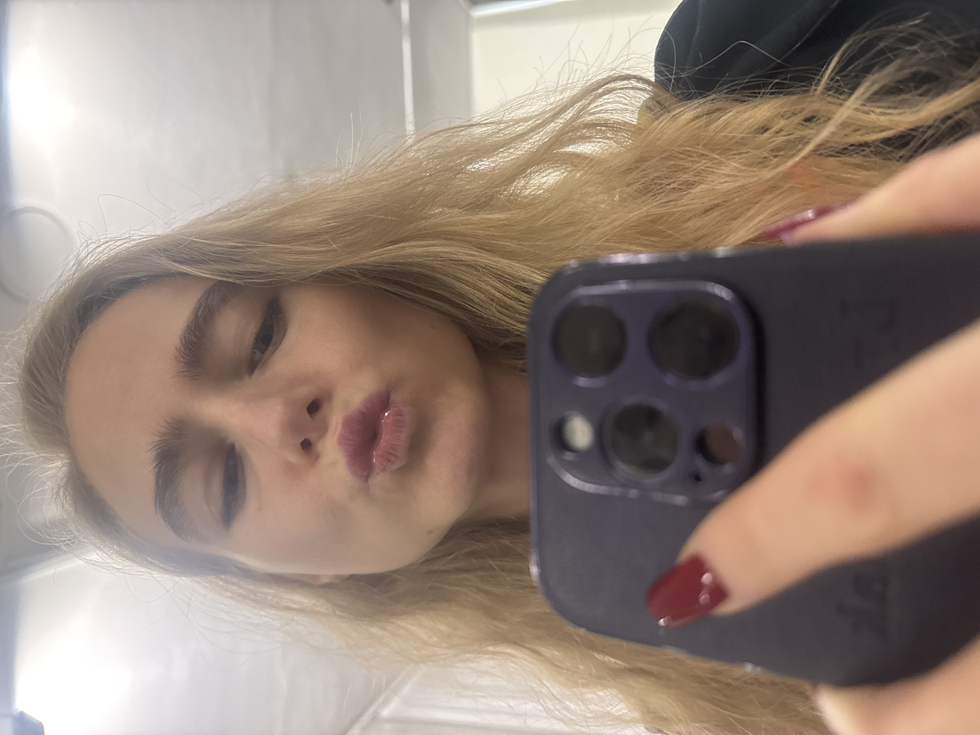 A close-up selfie captures a blonde female from the chest up, centrally positioned within an indoor environment in the city of Unknown, Unknown. Her head is slightly tilted, and her lips are puckered with glossy pinkish-red lipstick. She possesses full eyebrows and long, wavy blonde hair that frames her face. A dark top is partially visible on her right shoulder. Her right hand is visible in the lower foreground, holding a dark purple smartphone with a prominent multi-lens camera module on its rear. A finger with a dark red manicured nail extends over the phone. The background is brightly illuminated and appears as a light, non-descript reflective surface, consistent with an interior wall or ceiling.