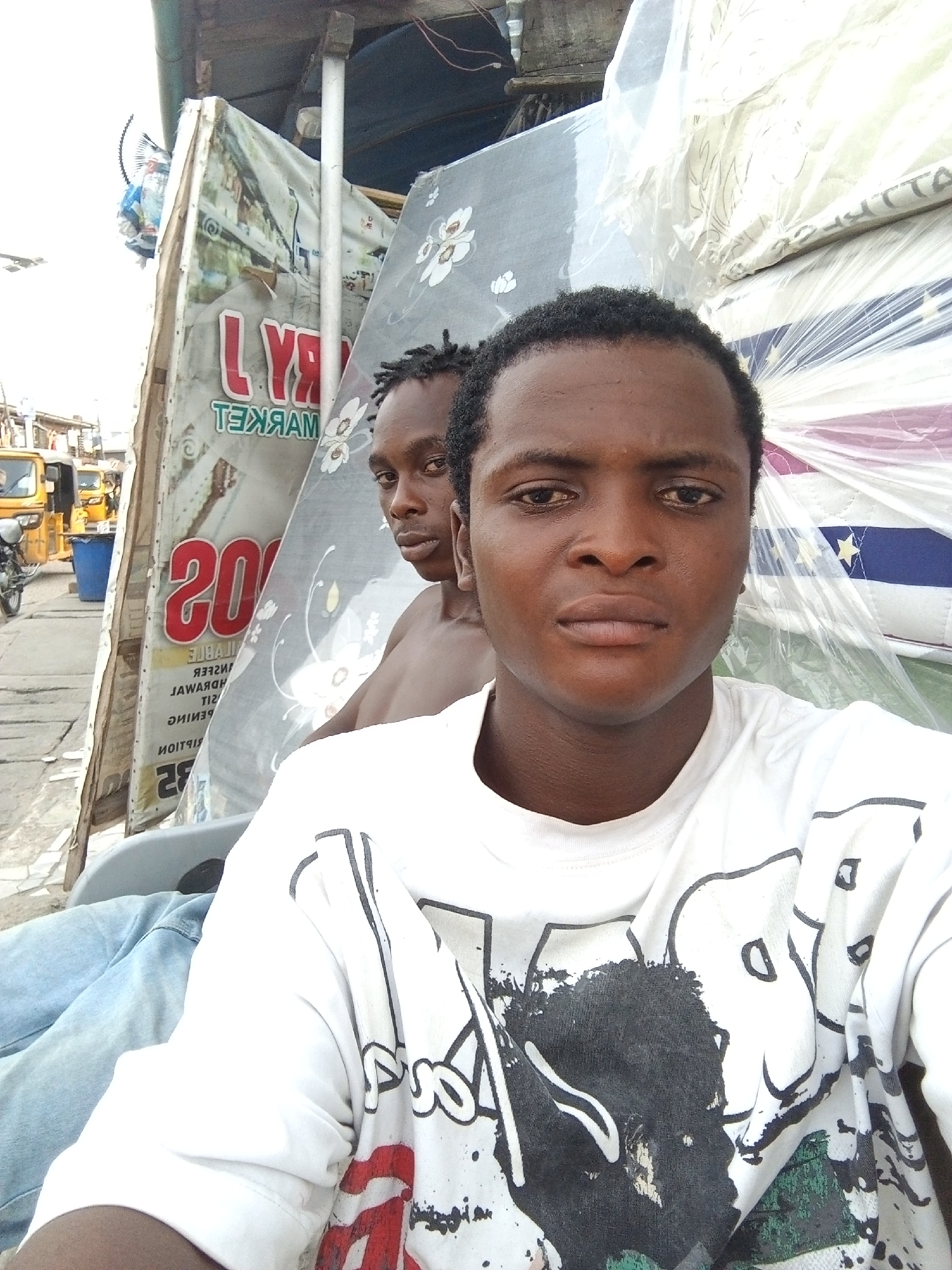 Two young men are captured in what appears to be a selfie taken outdoors in a bustling street or market area in Ado, Nigeria.
The man in the foreground is dark-skinned with short, dark curly hair and dark eyes, looking directly at the camera with a neutral expression. He wears a white short-sleeved t-shirt featuring a large black and red graphic design, possibly an abstract figure or face with paint splatters.
Behind him, slightly to his left, a second man with dark skin and short, dark hair (possibly dreadlocks or twists) looks back over his right shoulder towards the camera with a serious gaze. He appears to be shirtless.
The background is filled with elements of an outdoor market. To the right of the subjects, multiple mattresses are stacked, some covered in clear plastic, others displaying patterns like white flowers on a dark background, or blue and white stripes with yellow stars. Behind the men, a large, worn banner is visible on a building or stall structure. The text on this banner is reversed, clearly showing "LYRA MARKET" (ТЕХЯАМ АЯYL) in prominent red letters, with the number "200" also visible below it.
To the far left, a street scene unfolds under bright daylight, suggesting a sunny day. Yellow auto-rickshaws (locally known as Keke Napep), a blue plastic bin, and a motorcycle can be seen, further emphasizing the urban market setting. The overall impression is a candid shot within a vibrant, everyday scene.