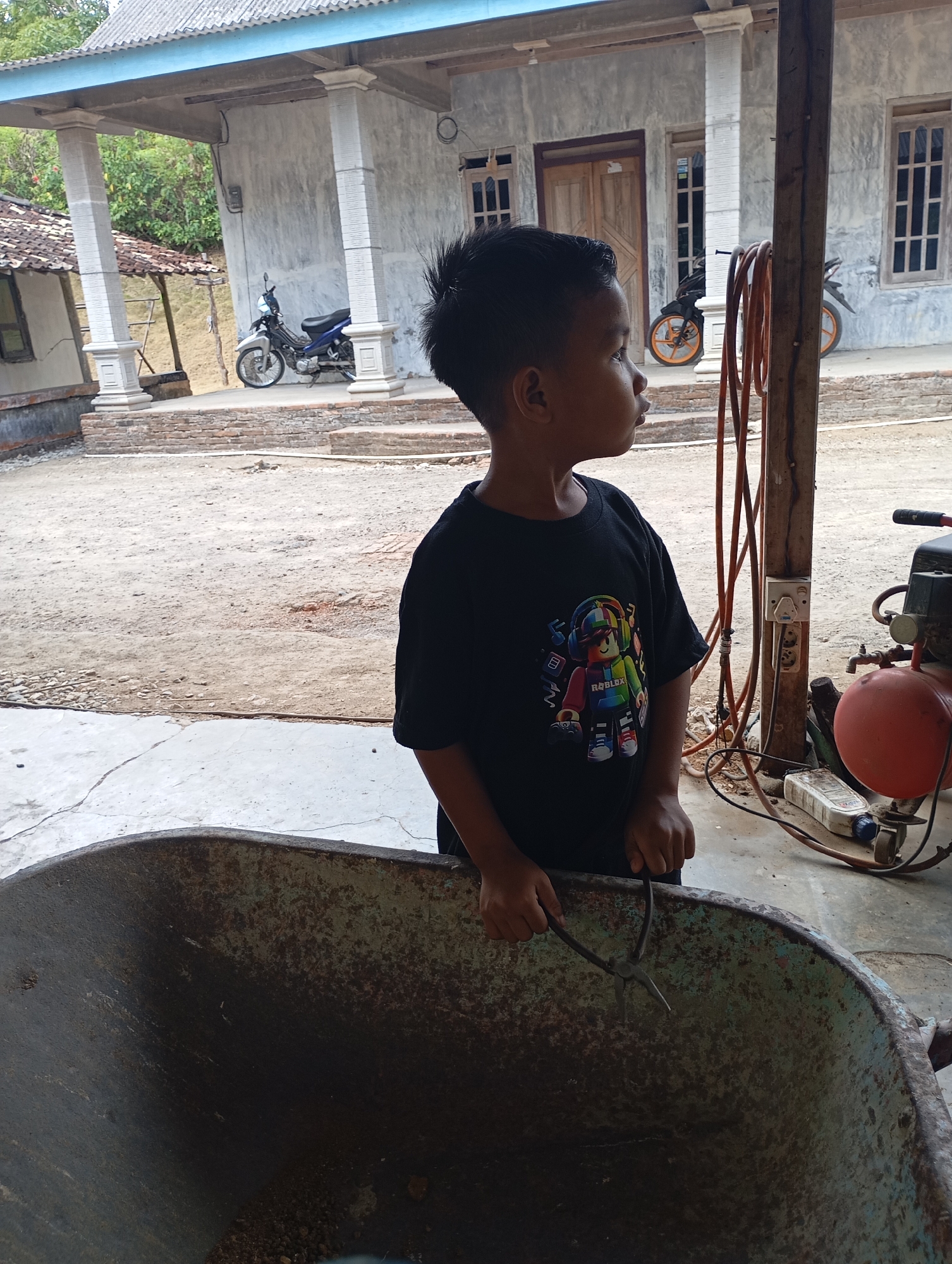 The image depicts a young boy standing next to a rusty wheelbarrow in what appears to be a residential yard in Ketepung, Indonesia. The boy, wearing a black t-shirt with a colorful graphic of a character from the game Roblox, is looking to his right. He is holding a pair of pliers in his hands. Behind him, a house with a gray concrete wall and a blue-trimmed roof is visible, with two motorcycles parked on the porch. The ground is covered in dirt and gravel. To the right of the boy, there is an air compressor and some electrical cords hanging from a wooden post. The overall impression is of a casual, everyday scene in a rural or semi-urban Indonesian setting.