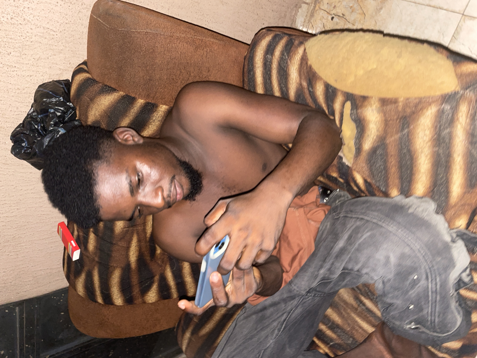 A young, shirtless Black man with a short haircut and a goatee is sitting in a chair with a tiger-striped pattern. He is wearing dark grey jeans. He is holding a blue cell phone in his hands and appears to be looking at it. A red and white package, possibly a cigarette pack, is on the armrest of the chair. A black plastic bag is on the back of the chair. The chair is placed in a room with a beige wall and tiled floor. The environment suggests an indoor setting, likely a residence, in Lokoja, Nigeria.