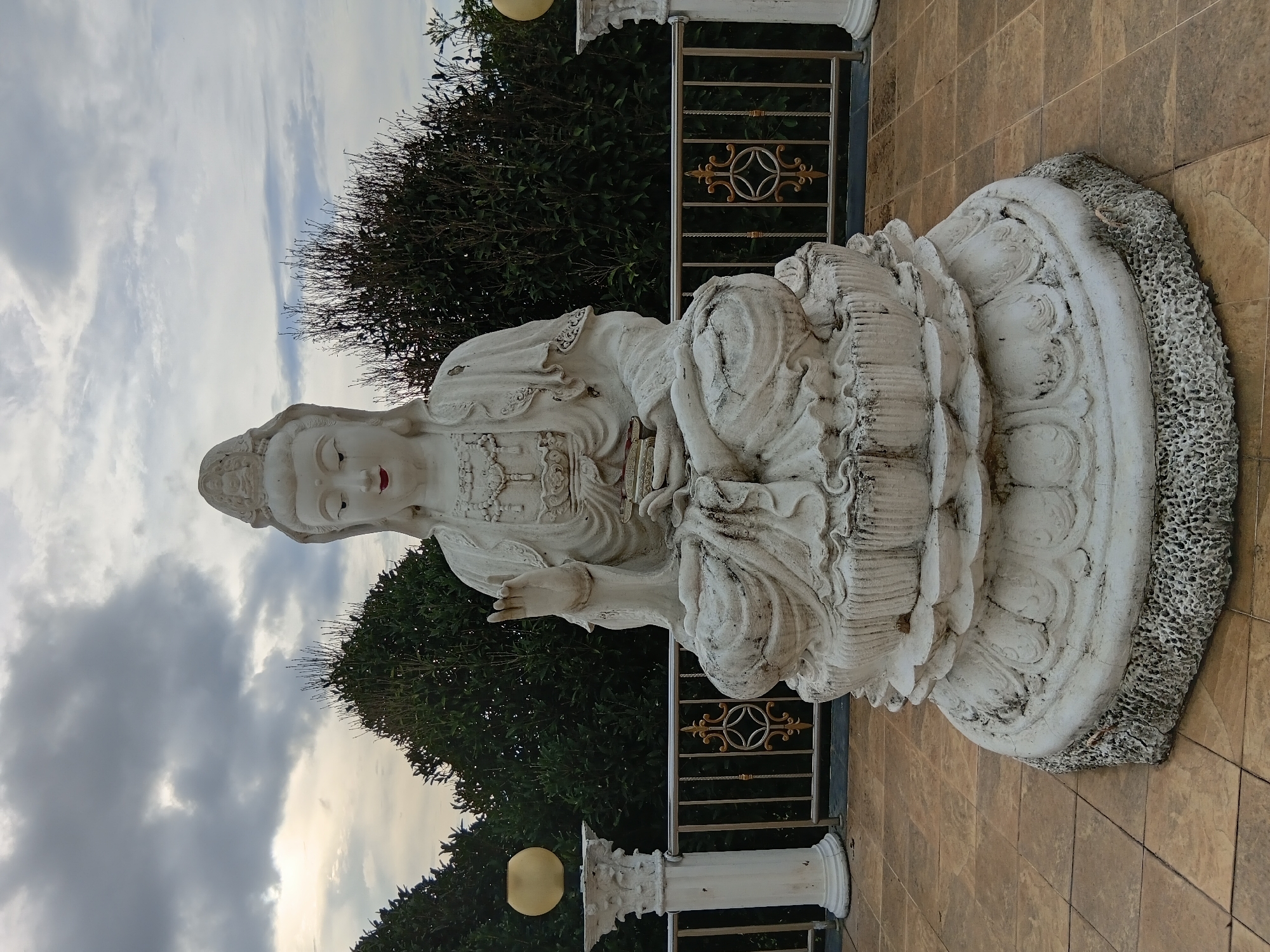 A white statue of a seated deity, possibly Guanyin, is positioned outdoors in front of a dense line of dark green trees. The statue, made of weathered white material, depicts a figure with serene facial features, red lips, and ornate headwear. It is seated on a multi-tiered lotus pedestal. The deity's right hand is raised in a gesture of blessing or protection, while the left hand rests in its lap, holding a small object.
The statue is placed on a tiled patio area, which appears to be part of a temple or religious site. A metal railing with decorative gold accents separates the patio from the trees behind. On either side of the statue, white pillars topped with yellow, spherical lamps are visible. The sky above is overcast with a mix of dark and lighter clouds, suggesting a cloudy day.
The location is identified as Ban Lao Phatthana, Thailand. The overall scene conveys a sense of peace and reverence.