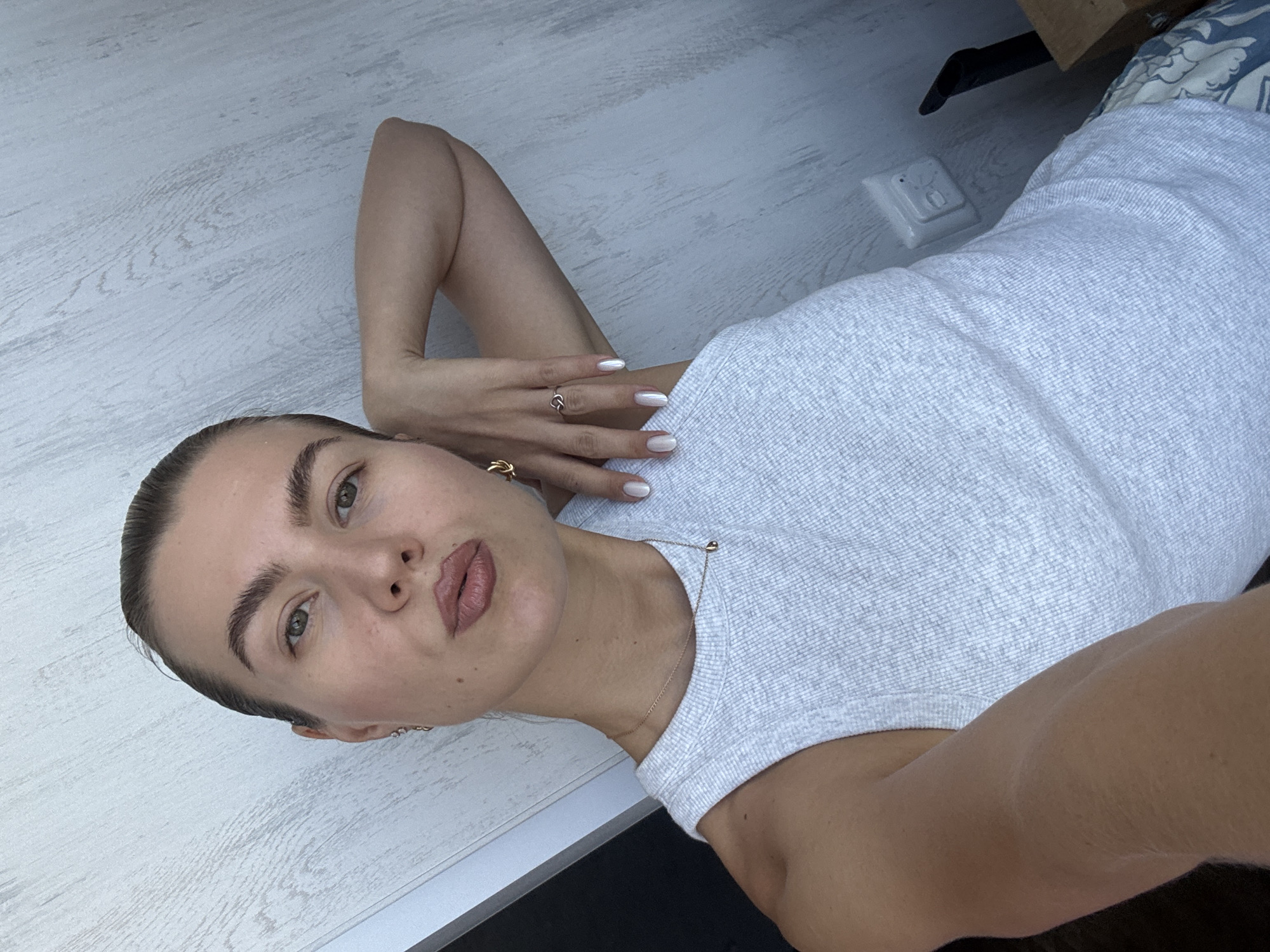 In a close-up, selfie-style shot, a woman with her hair pulled back leans against a textured, pale wood-paneled wall. She wears a light gray, ribbed, sleeveless tank top and delicate gold jewelry, including a thin chain necklace with a teardrop pendant, small hoop earrings, and two rings on her right hand. Her left arm is bent upwards, resting behind her head, while her right hand is placed on her right hip. Her nails are painted a pearlescent white. Her makeup includes subtle eyeliner and lip gloss. A white electrical outlet is visible on the wall to her right. The lighting suggests it is daytime.