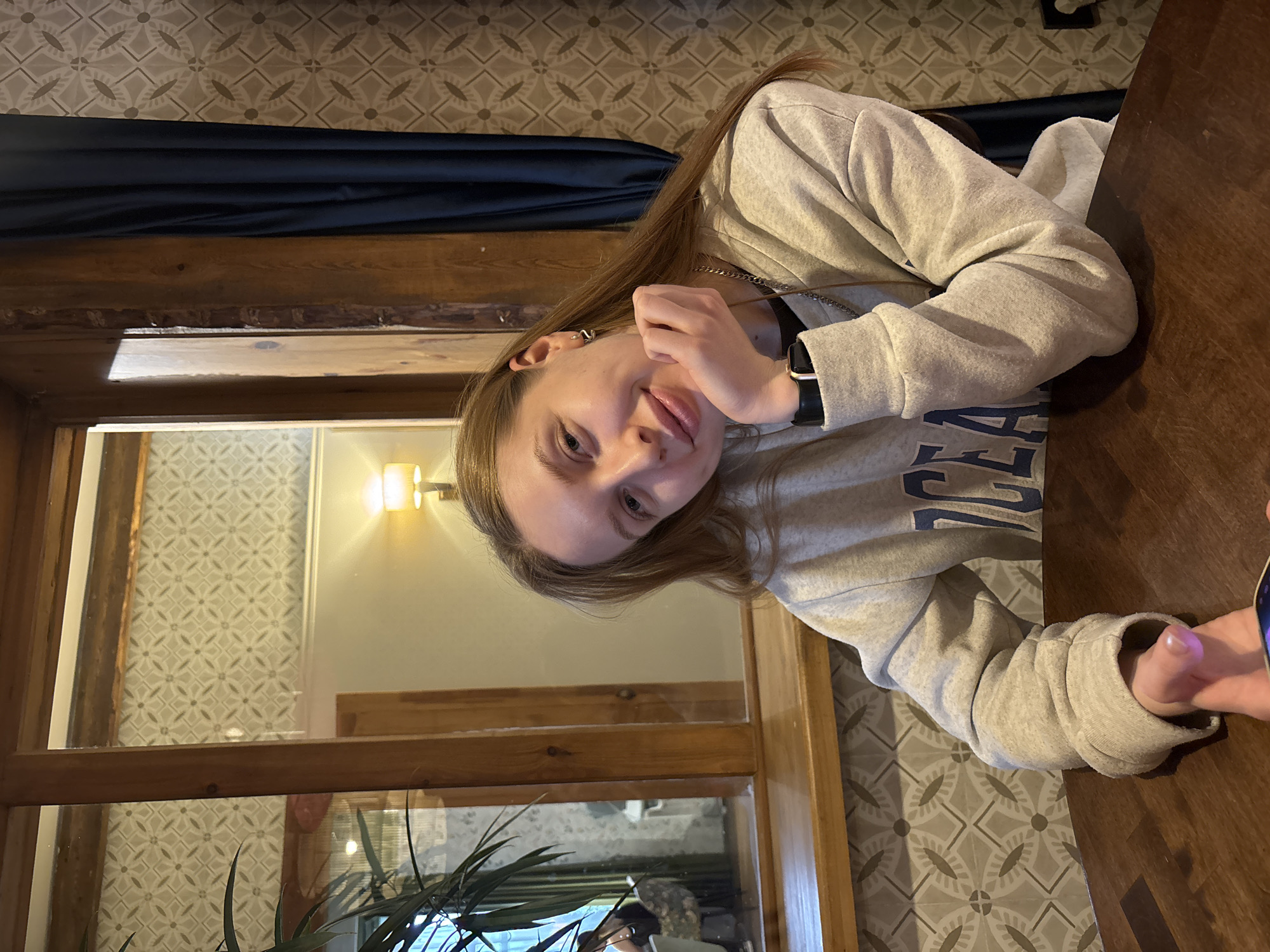 A young woman is sitting at a wooden table in what appears to be a restaurant or cafe. She is wearing a gray sweatshirt with blue lettering and a silver chain necklace. Her left hand is raised near her face. She has light skin, brown hair, and is looking towards the camera. Behind her, a window frames a view of a room with patterned wallpaper and a lit wall lamp. The location is Perm, Russia, as indicated by the metadata associated with the image.