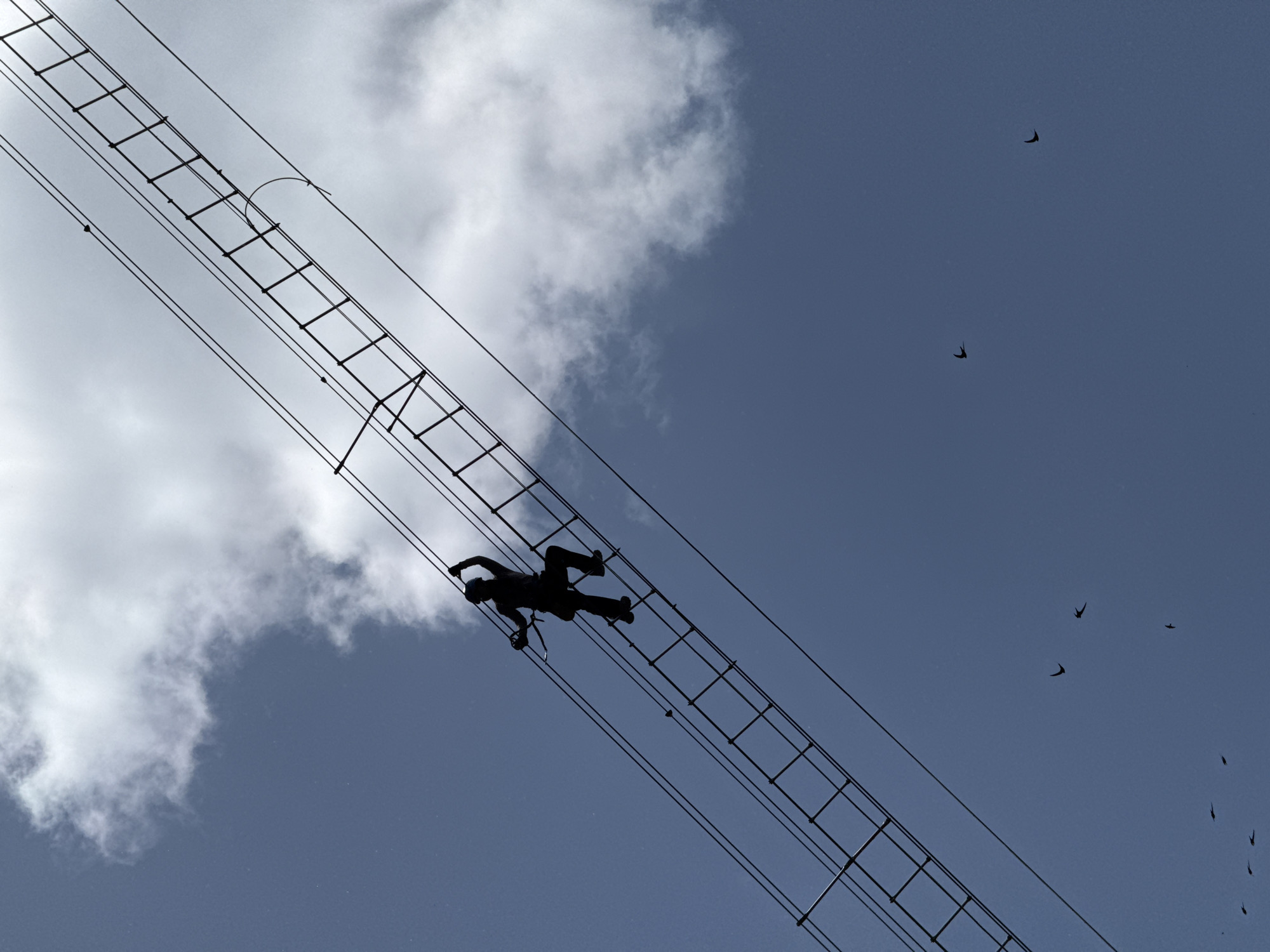 A low-angle shot shows a silhouetted figure on a suspension bridge against a backdrop of clouds and a blue sky in Yongding District, China. The figure appears to be lying face-down on the bridge's ladder-like structure. Several birds are silhouetted against the blue sky.
