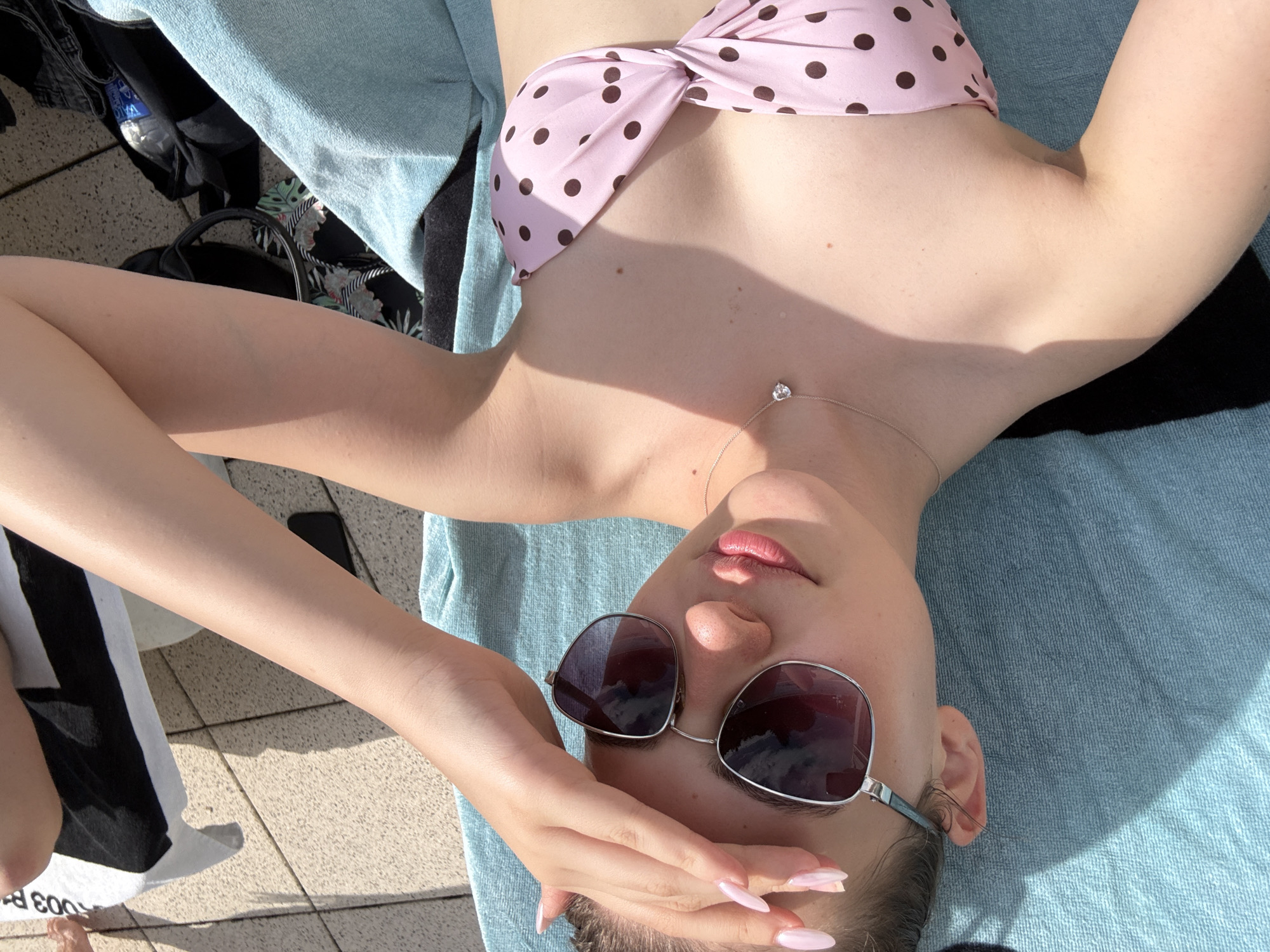 A young woman with light skin and dark hair pulled back is lying down on a blue towel, presumably outdoors near a swimming pool, indicated by the tiled ground and shadows. She is wearing a pink bikini with brown polka dots and a silver necklace with a heart-shaped pendant. She has sunglasses on and is touching her forehead with her right hand. The lighting suggests a sunny day. The background includes a black and white striped towel and a portion of another person's leg is visible. The scene is in Forbach, Germany.