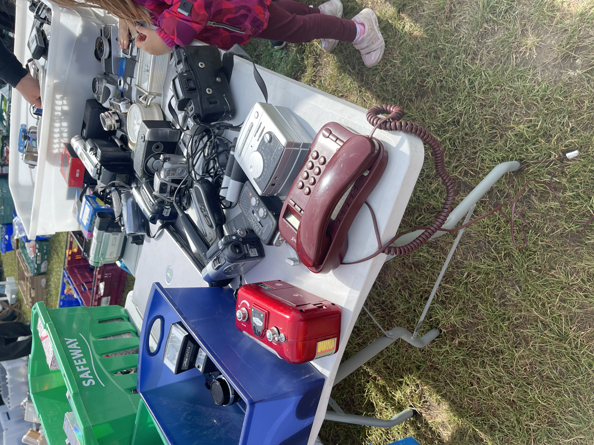 The image depicts a table covered with assorted electronic items, seemingly at an outdoor market or sale. A child is seen examining items on the table. The table displays a variety of devices, including cameras, radios, and a landline telephone. Below the table, crates are used for storage. The location appears to be a grassy area, possibly in the vicinity of Sevenoaks, United Kingdom. A green crate labeled "SAFEWAY" is visible. The objects suggest a collection of vintage or used electronics.