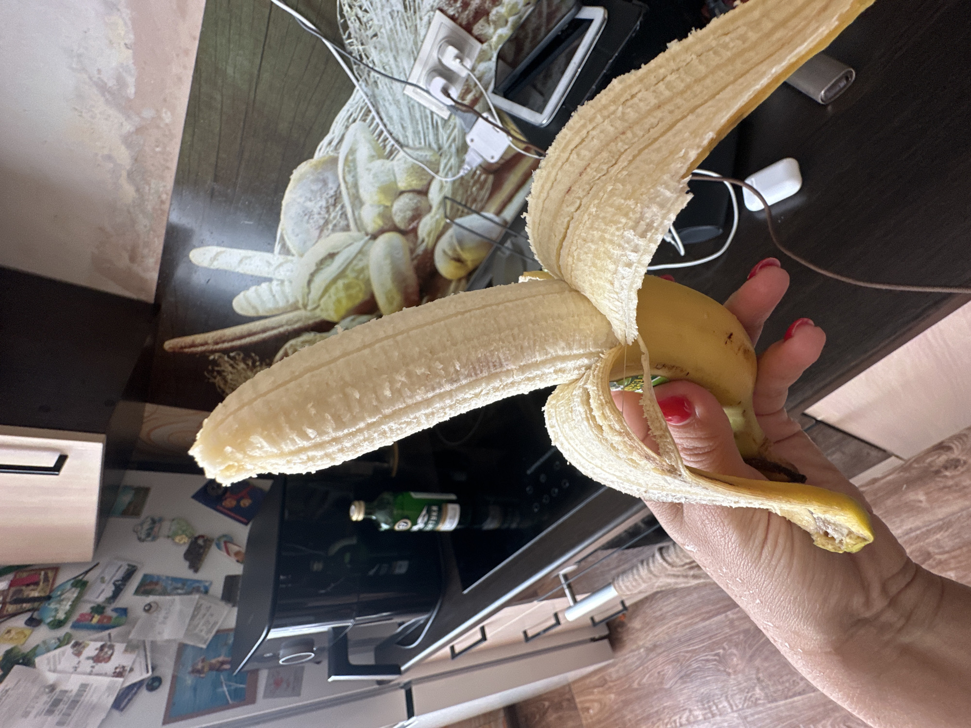 An unpeeled banana is held in a person's hand, with a portion of the peel peeled back. The background shows a kitchen counter with various items, including an air fryer, a bottle of liquid, and electrical outlets with cords. The floor is made of wooden planks. The image is a close-up of the banana and hand, with the kitchen environment blurred in the background. The location is a kitchen in Novoaltaysk, Russia.