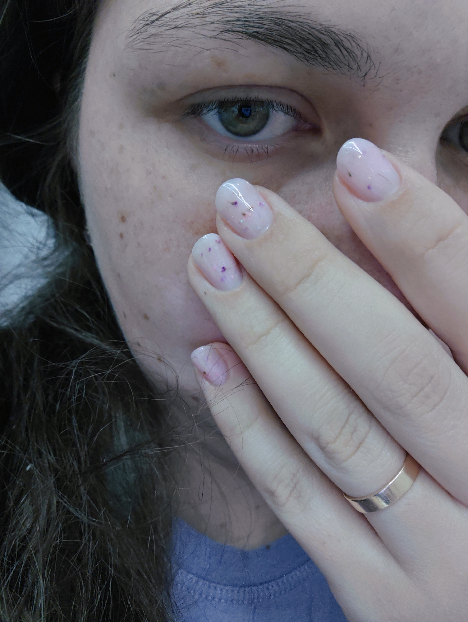A close-up portrait of a woman with fair skin, characterized by freckles on her face, especially around the cheek and nose. She has a single visible green eye. Her hand is held up near her face, obscuring part of it. Her nails are painted a light pink color with small purple and silver flecks. She is wearing a plain gold ring on one finger. Strands of brown hair are visible near her ear and in the lower-left corner of the image. Part of a light purple or blue garment is seen in the lower part of the frame. The image does not contain sufficient information to determine the location as Chaikovsky, Russia, or any specific environment.
