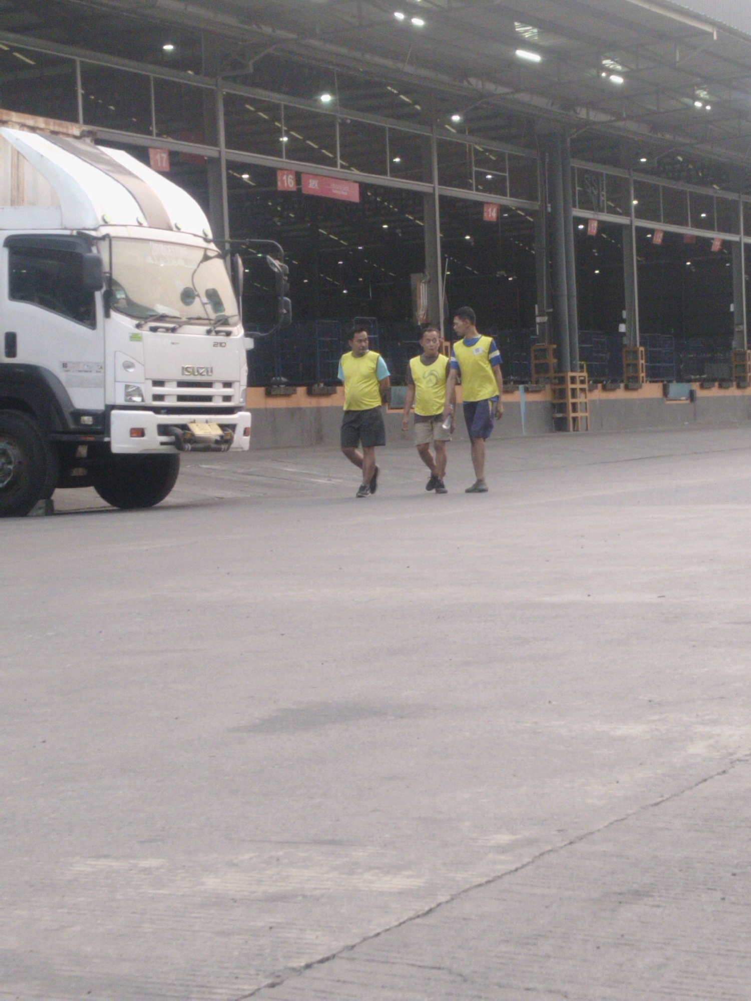 In Belimbing, Indonesia, an industrial area under a covered loading dock with overhead lighting features a white Isuzu truck. Three men are walking across a paved surface. Two men, wearing yellow vests over short-sleeved shirts and shorts, walk together in the middle ground. One man on the right, also wearing a yellow vest over a blue shirt and dark shorts, walks separately towards the right foreground. The men appear to be engaged in casual conversation. The environment suggests a logistics or distribution center.