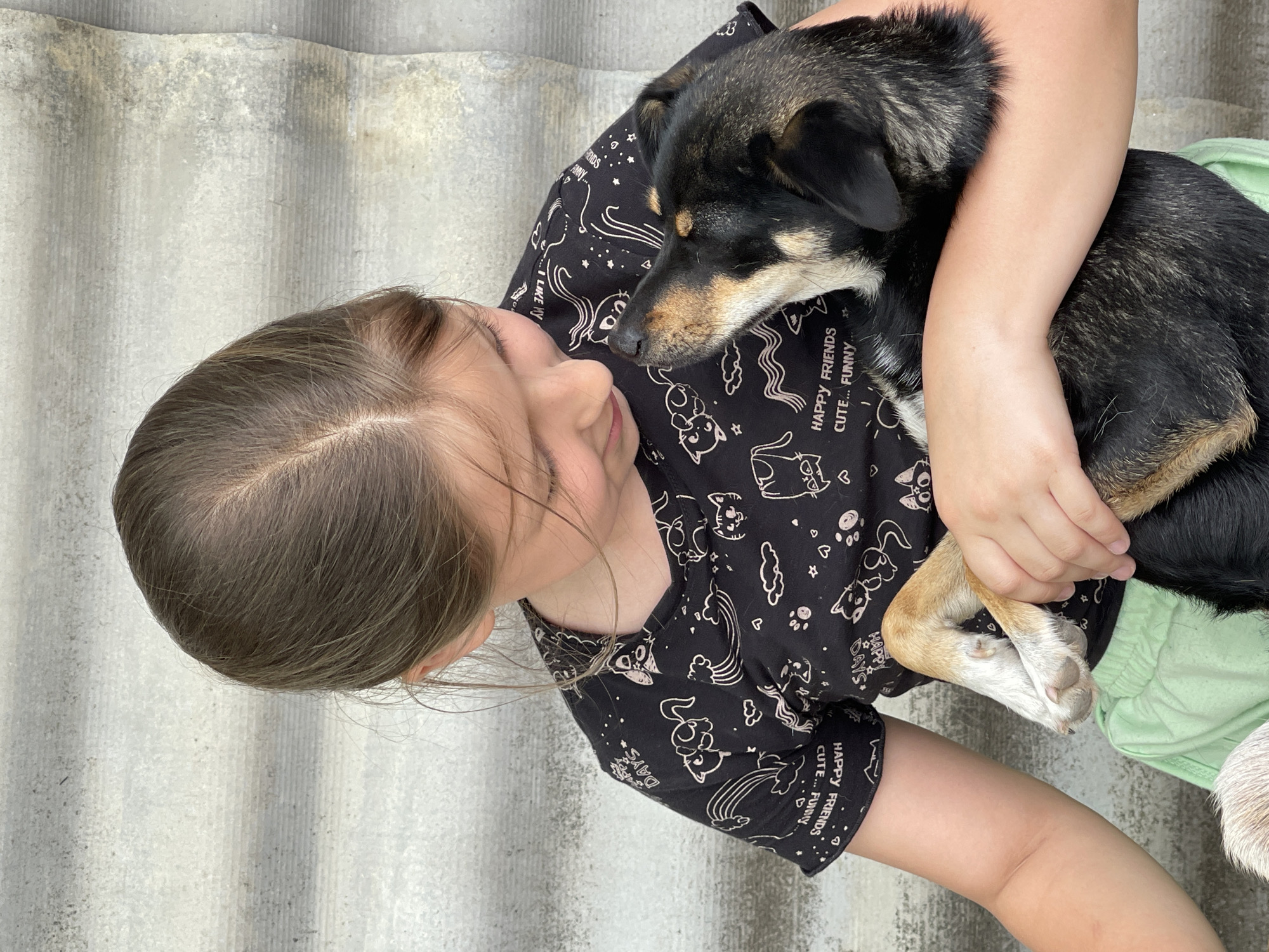 An outdoor medium shot captures a young girl with brown hair, wearing a black t-shirt with a white cartoon print and light green shorts, embracing a black and tan dog. The girl, looking down and to the left, gently holds the dog, which is looking up at her. The background consists of a gray, corrugated wall. The scene appears to be located in the village of Levokumka, Russia.