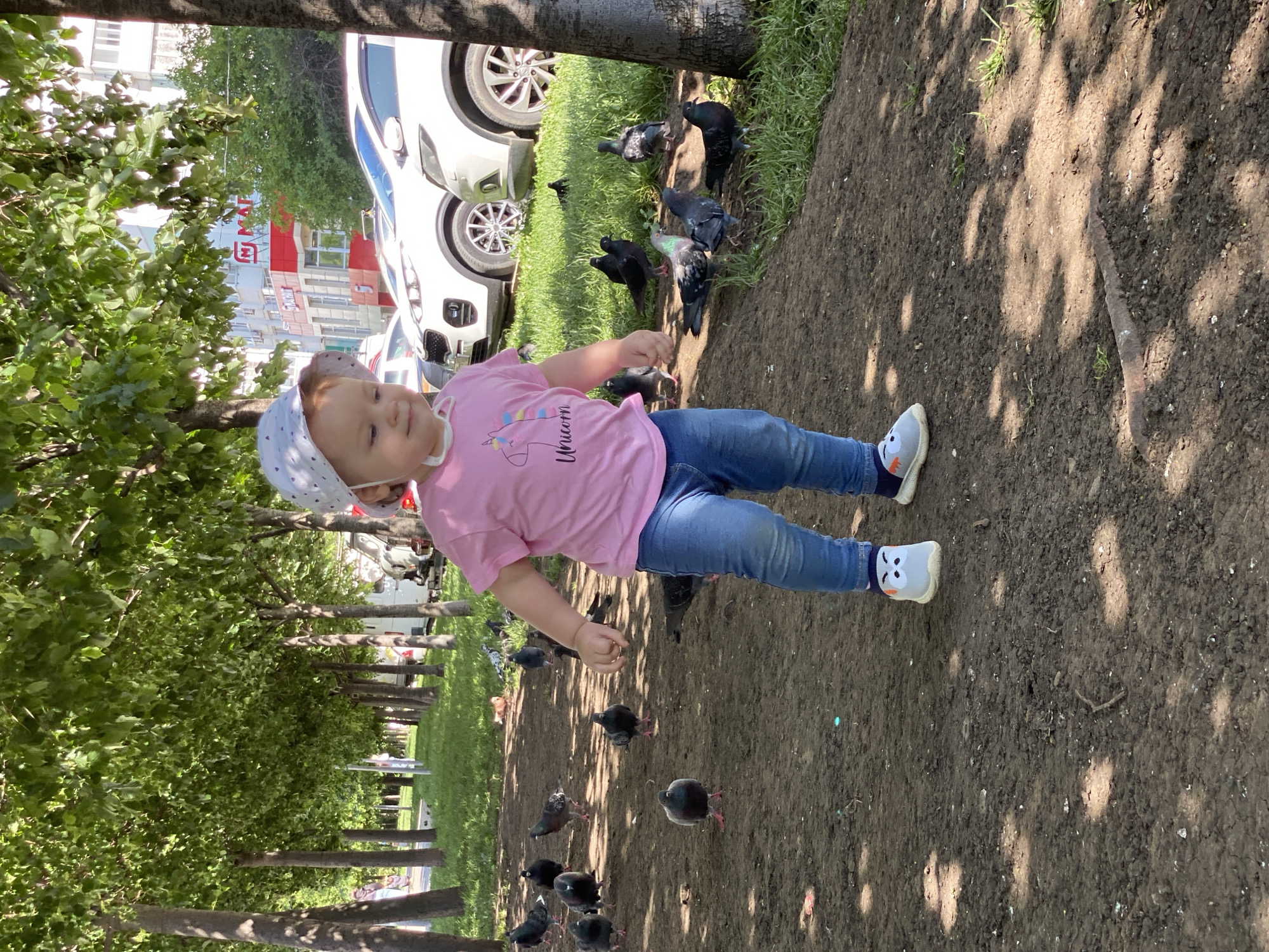 A baby girl in a pink unicorn shirt and blue jeans stands on a dirt ground, surrounded by pigeons in a park. She is wearing a white bandana and patterned socks with gray shoes. She appears to be feeding the pigeons. Trees line the park's edge. A white car is parked nearby. Buildings are visible in the background. The location is Unknown, Unknown.
