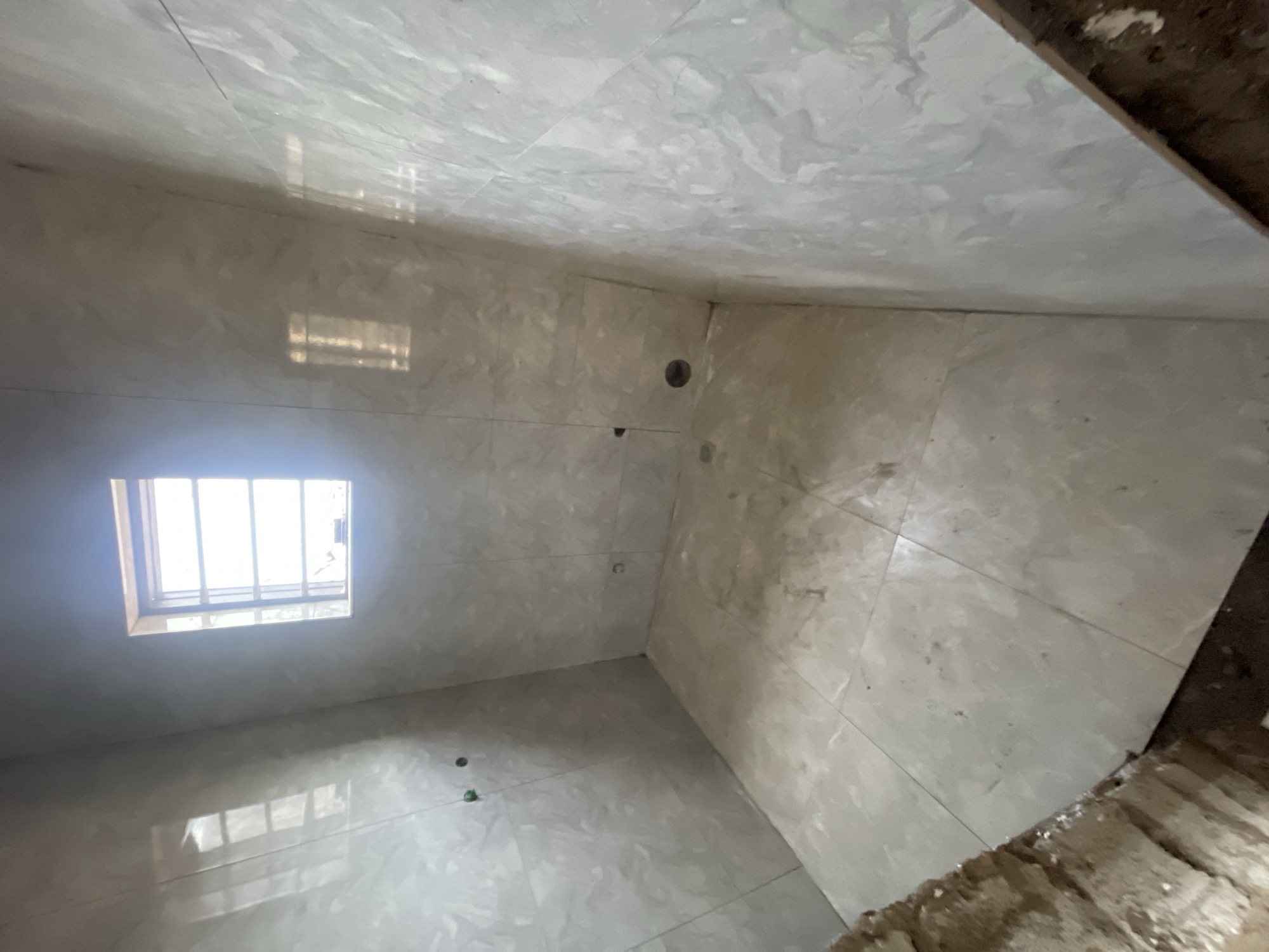 The image shows the interior of a small, unfinished bathroom. The walls, floor, and ceiling are covered in large, light-colored marble tiles. There is a window with bars on the left wall, and the sunlight coming through is reflecting off the tiled floor. There are a few holes in the walls, possibly for plumbing or fixtures. The floor near the edge of the room on the right is unfinished, showing what looks like concrete or brick. The location is Angwan Sayawa, Nigeria.