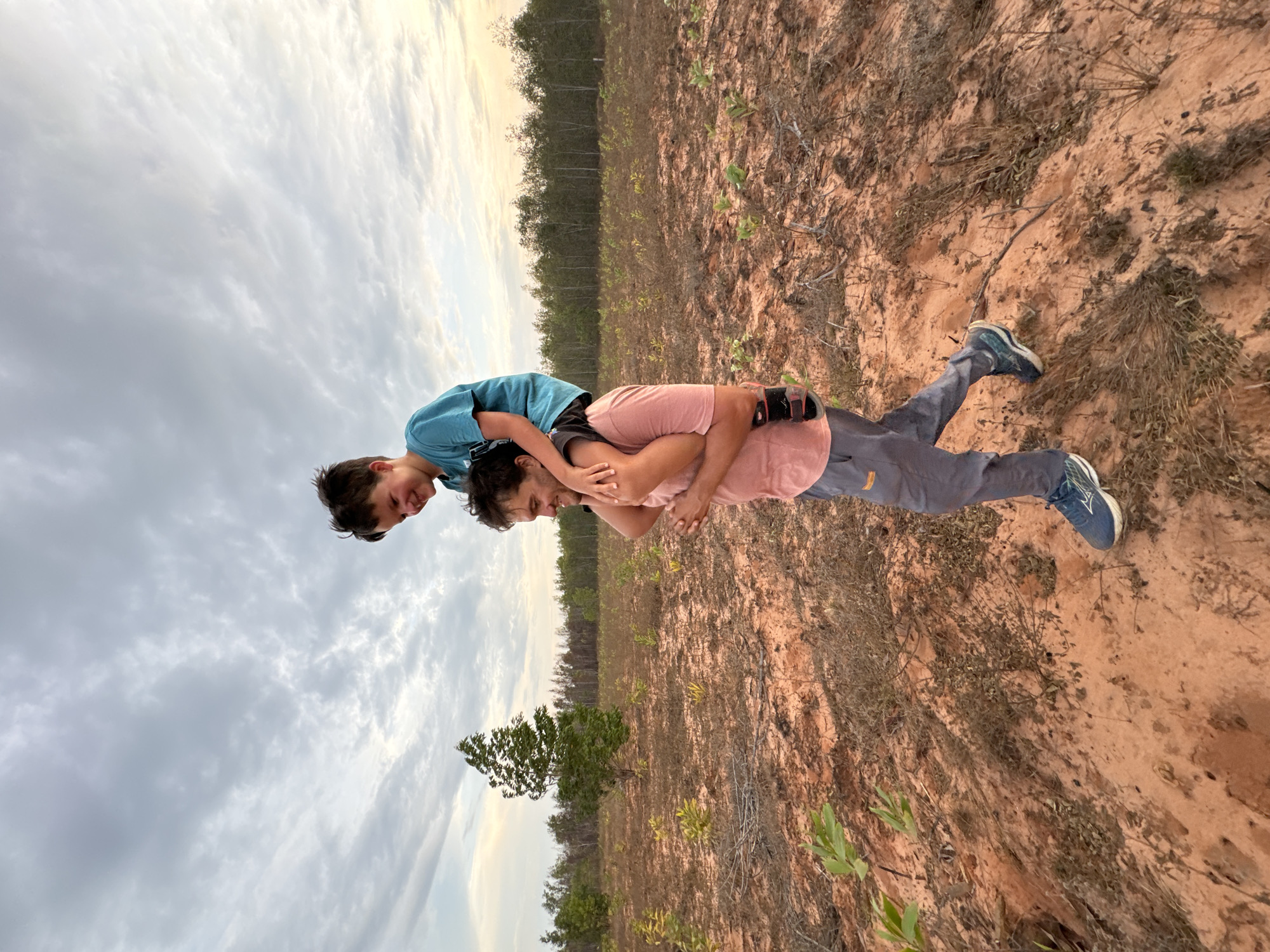 Two people are in a field in Phu Thuy Ward, Vietnam. One person is giving the other a piggyback ride. The person on top is wearing a blue shirt, and the person on the bottom is wearing a salmon-colored shirt. The field is dirt and sparse vegetation. The sky is cloudy.