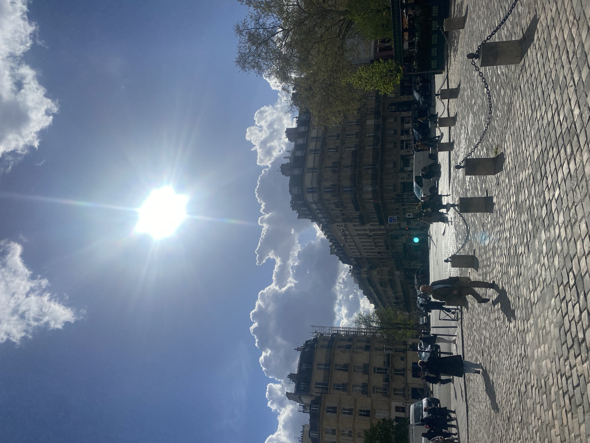 The image captures a sunny daytime street scene at Place Saint-Michel in Paris, France, viewed from the southern side, looking generally north-east. The sky is bright blue with prominent, voluminous white cumulus clouds. The sun is positioned in the upper center-right, casting a strong starburst lens flare and long, distinct shadows across the scene.
The foreground and mid-ground are paved with light-colored cobblestones (pavé). A series of six dark grey stone bollards, connected by heavy metal chains, delineates an area from the lower center-right extending diagonally towards the mid-left.
Numerous pedestrians are visible. In the mid-ground, a person wearing a light brown jacket and trousers walks from right to left, casting an elongated shadow. Another individual, dressed in a dark coat, walks away from the camera towards the left. Additional pedestrians are scattered across the sidewalks and crossing areas in the background.
The surrounding architecture consists of multi-story, classic Parisian Haussmannian buildings with stone facades, balconies, and dormer windows. A prominent building on the far left features a rounded corner. Further down the street on the left, a building shows extensive scaffolding along its side.
In the background, several vehicles, including a white van and dark-colored cars, are present on the streets, indicating active traffic. A green traffic signal is visible on the right side of the main intersection. On the right, large trees with light green foliage suggest a spring season, partially obscuring a building with dark awnings, possibly indicating an outdoor cafe.