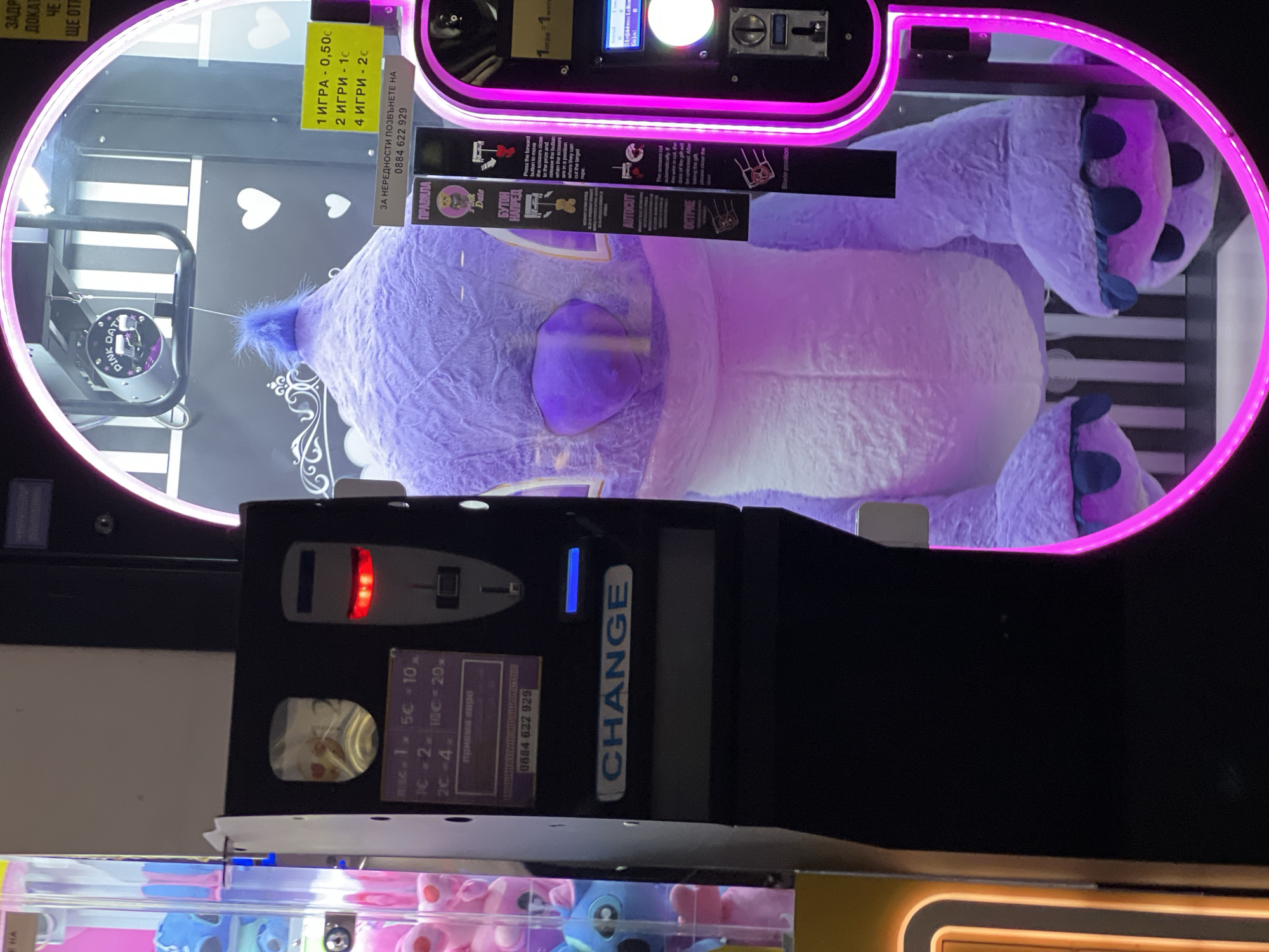 A front-facing, eye-level medium shot shows a plush purple stuffed animal in a claw machine. The plush has a large, rounded head, floppy ears, and a blue tuft of hair. The machine's front panel is black with an inset coin slot area, a "CHANGE" label in blue lettering, and instructions in Bulgarian. Red and blue indicator lights flank the coin slot. The play area has a pink neon border and a black and white striped background. Above the stuffed animal, signs in Bulgarian detail pricing (1 play - €0.50, 2 plays - €1, 4 plays - €2) and contact information for complaints. The claw machine's interior contains the cutting mechanism and detailed instructions for use with diagrams. Below the machine, other plush animals are visible. The scene is likely in an arcade or entertainment area in Plovdiv, Bulgaria.