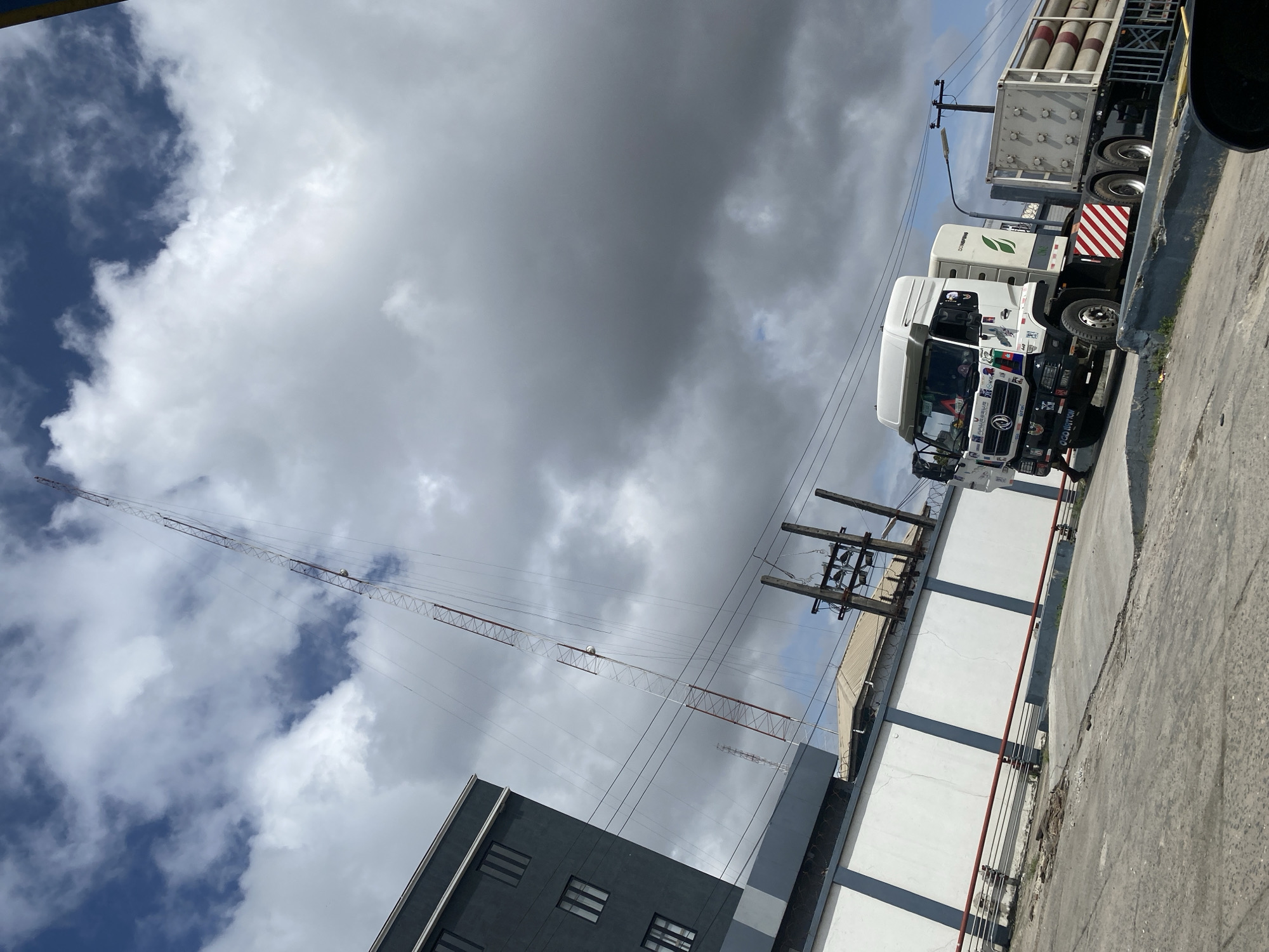 In Ajah, Nigeria, a white truck with a trailer carrying cylindrical objects is parked on an inclined concrete surface. The truck is adorned with various stickers and decals, including one that reads "OGD NATION." To the left of the truck, a tall, slender communication tower with red and white latticework stretches into a cloudy sky. Adjacent to the tower is a multi-story dark grey building with visible windows. A white wall with dark grey vertical stripes runs along the middle ground of the scene, with a red railing along its base. Overhead power lines and utility poles are present. The ground appears to be a mixture of concrete and rough asphalt, with some debris visible. The overall environment suggests an industrial or commercial area within the city.