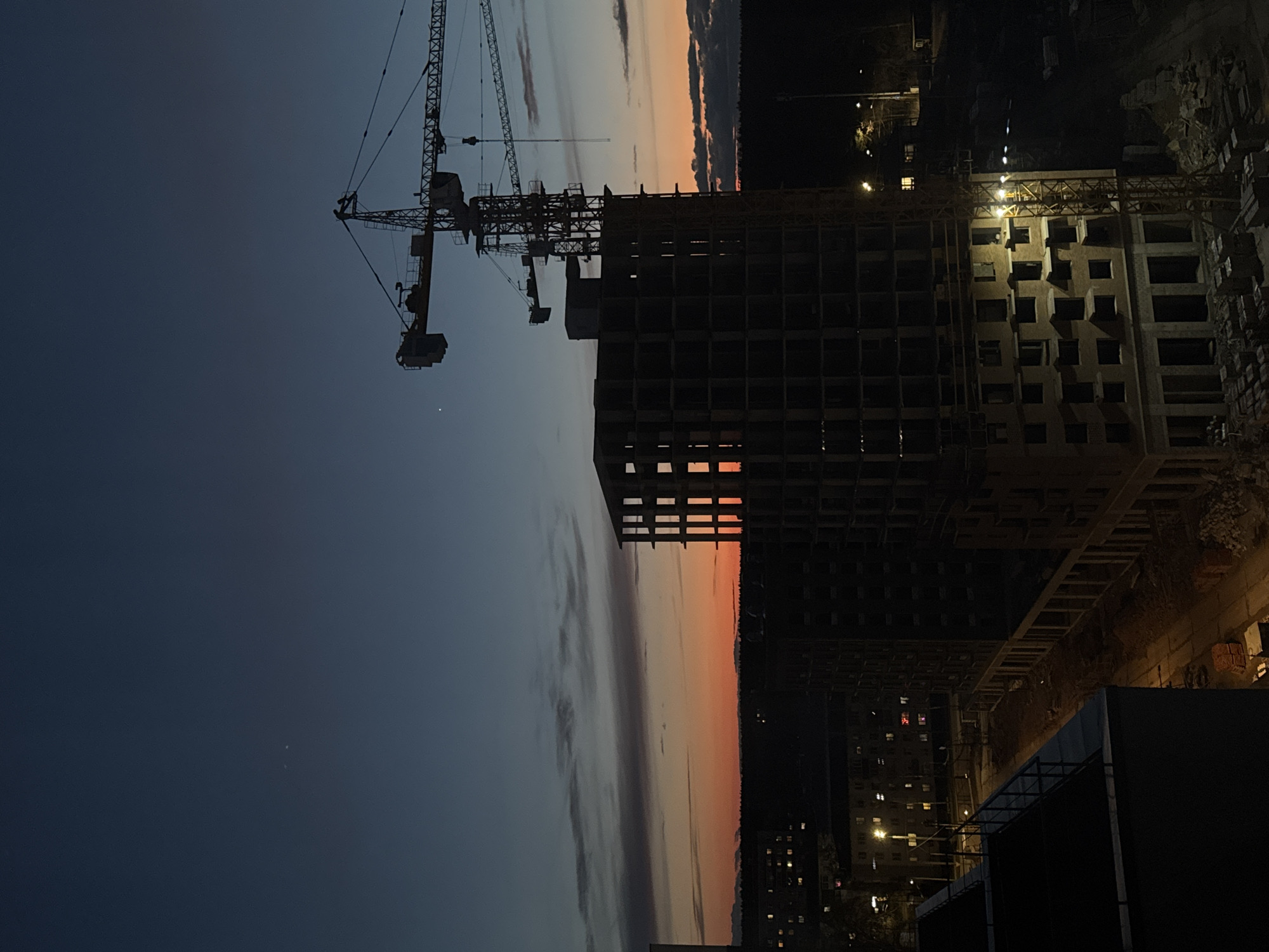The image, originally rotated ninety degrees, captures an urban construction site at dusk or sunset. In the foreground and mid-ground, the primary subjects are the dark silhouettes of two tower cranes and a large, multi-story building under construction.
The main tower crane dominates the left side, its skeletal structure and long arm extending across the frame. A second, less distinct crane is partially visible behind it. To the right, the unfinished building stands as a concrete shell, displaying numerous empty square openings for future windows and rooms. Several lights illuminate the building's upper levels, suggesting ongoing work, and some individual windows glow from within the lower floors.
The sky above presents a striking gradient, transitioning from a deep indigo at the top to a vibrant band of orange and red hues along the horizon, indicative of the setting sun. Streaky, dark clouds are visible across the mid-section of the sky. Below the colorful horizon, a dark urban landscape stretches into the distance, dotted with countless small, glowing lights from distant buildings, suggesting a bustling city (Izhevsk, Russia) coming to life at night. There is no visible text in the image.