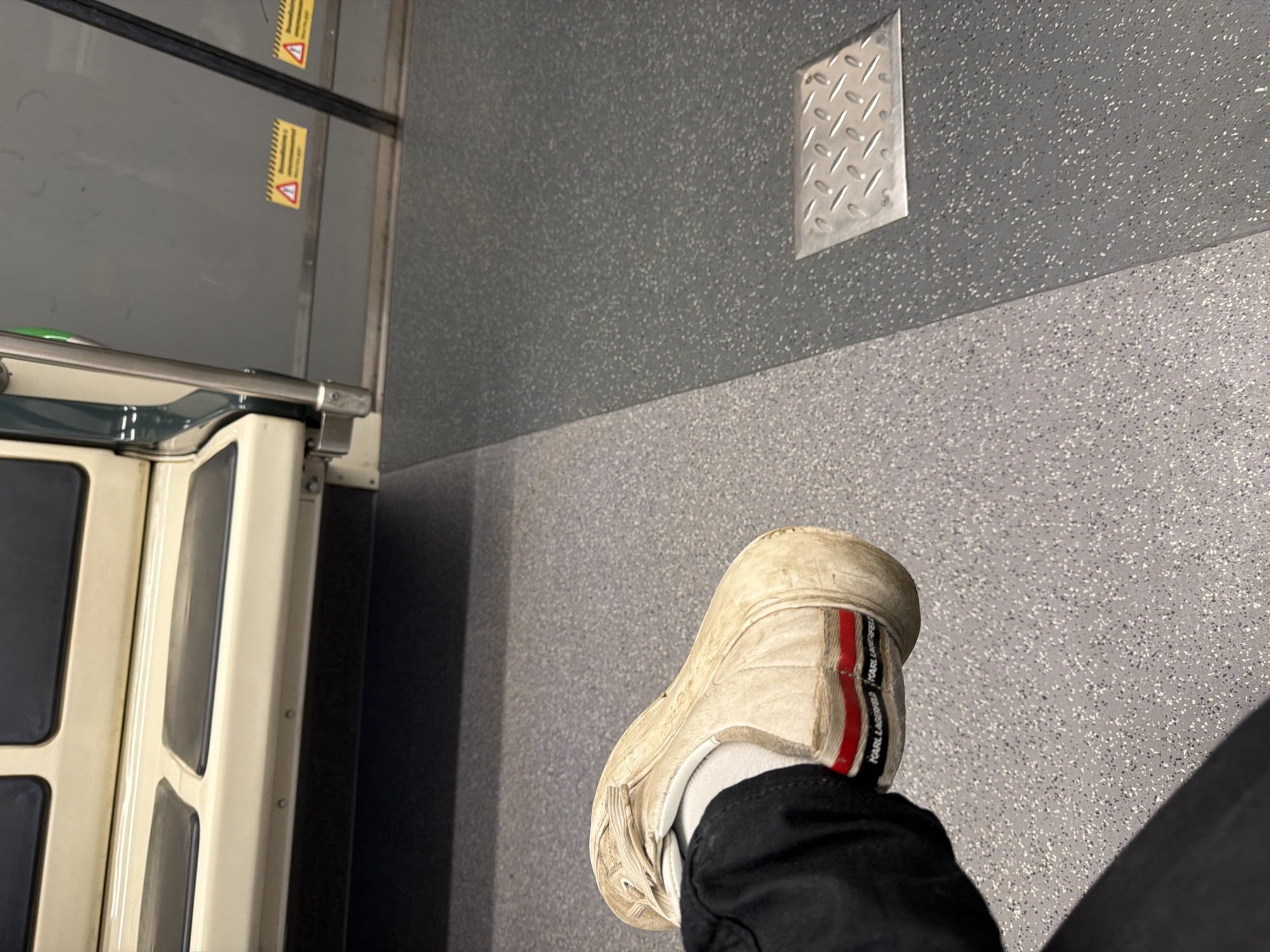 The image depicts the interior of public transportation, likely a bus or tram in Sofia, Bulgaria. A person wearing black pants and a white Karl Lagerfeld sneaker sits with their leg extended into the aisle. The floor and lower walls are gray with a speckled pattern. A metal, diamond-patterned step guard is affixed to the wall. To the left, a portion of a vehicle door with small windows is visible, along with yellow warning stickers.