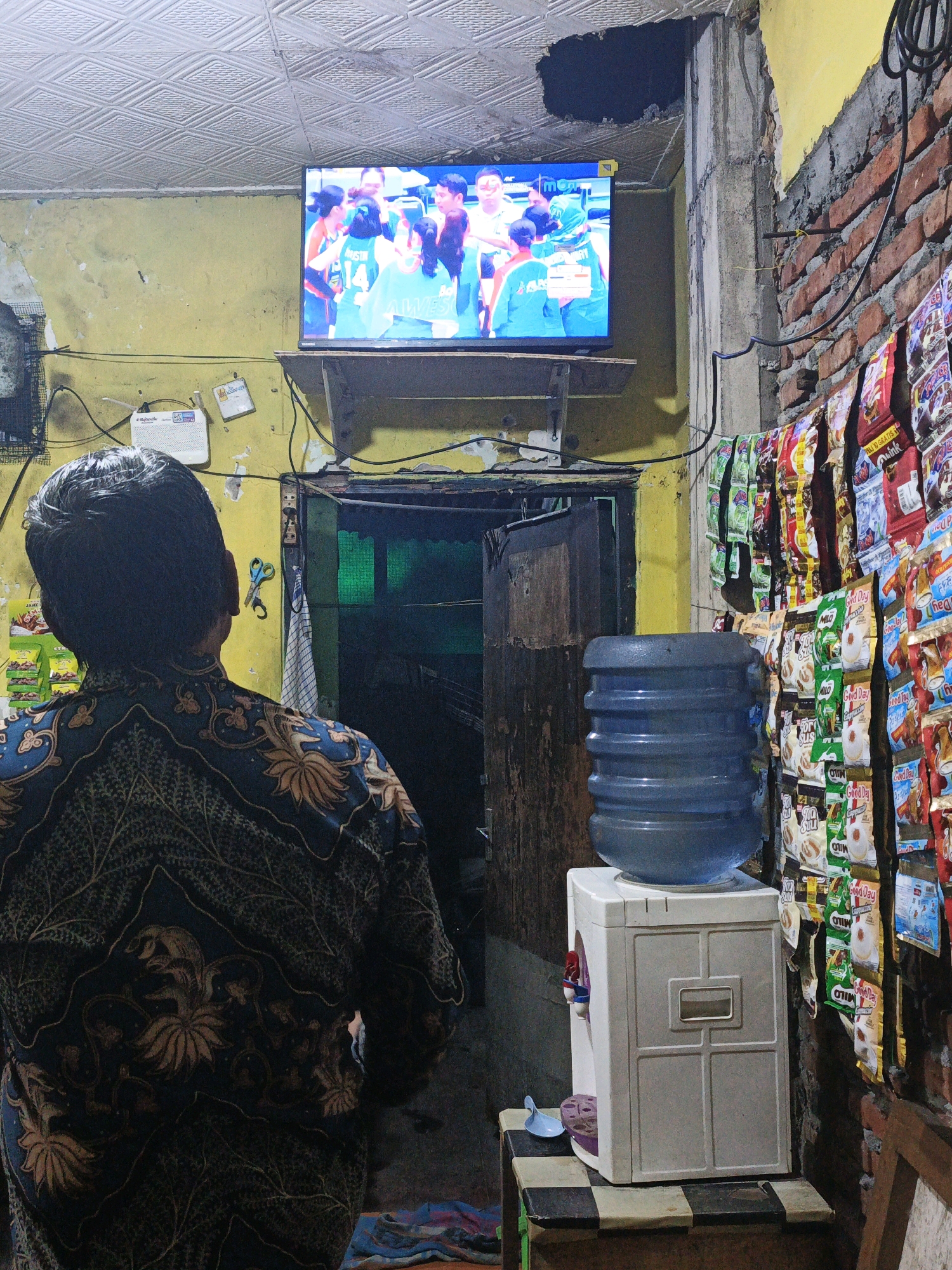 The image depicts an indoor scene in Ketegan, Indonesia, likely a modest dwelling or shop. A person, viewed from behind, wearing a dark blue batik shirt with a floral pattern, is watching a television screen mounted on the wall. The television displays a group of individuals in green athletic attire, possibly a sports team. The room is sparsely furnished and shows signs of wear and tear. The walls are painted yellow and exposed brick, with a textured ceiling. To the right of the television, a water dispenser with a large blue water jug sits on a white cooler. The wall beside the dispenser is adorned with numerous small packages of snacks or instant drinks hanging in rows. A doorway leading to another area is visible behind the water dispenser. Various wires and electrical outlets are present on the walls, suggesting a functional but not necessarily tidy living space. The overall impression is of a lived-in, practical environment.