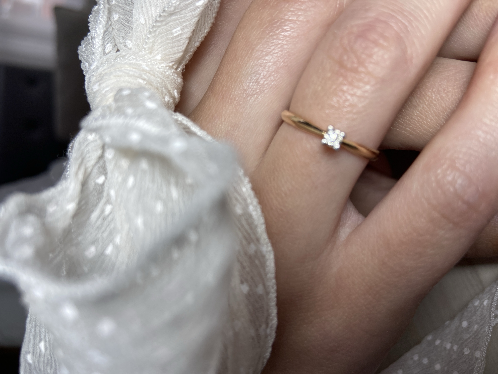 A close-up shot of a person's hand wearing a gold ring with a small, possibly square-cut diamond. The ring is on the fourth finger of the hand. Part of a white, textured fabric with small dots is visible to the left and below the hand. The image lacks specific details that would indicate the location as Budva, Montenegro.