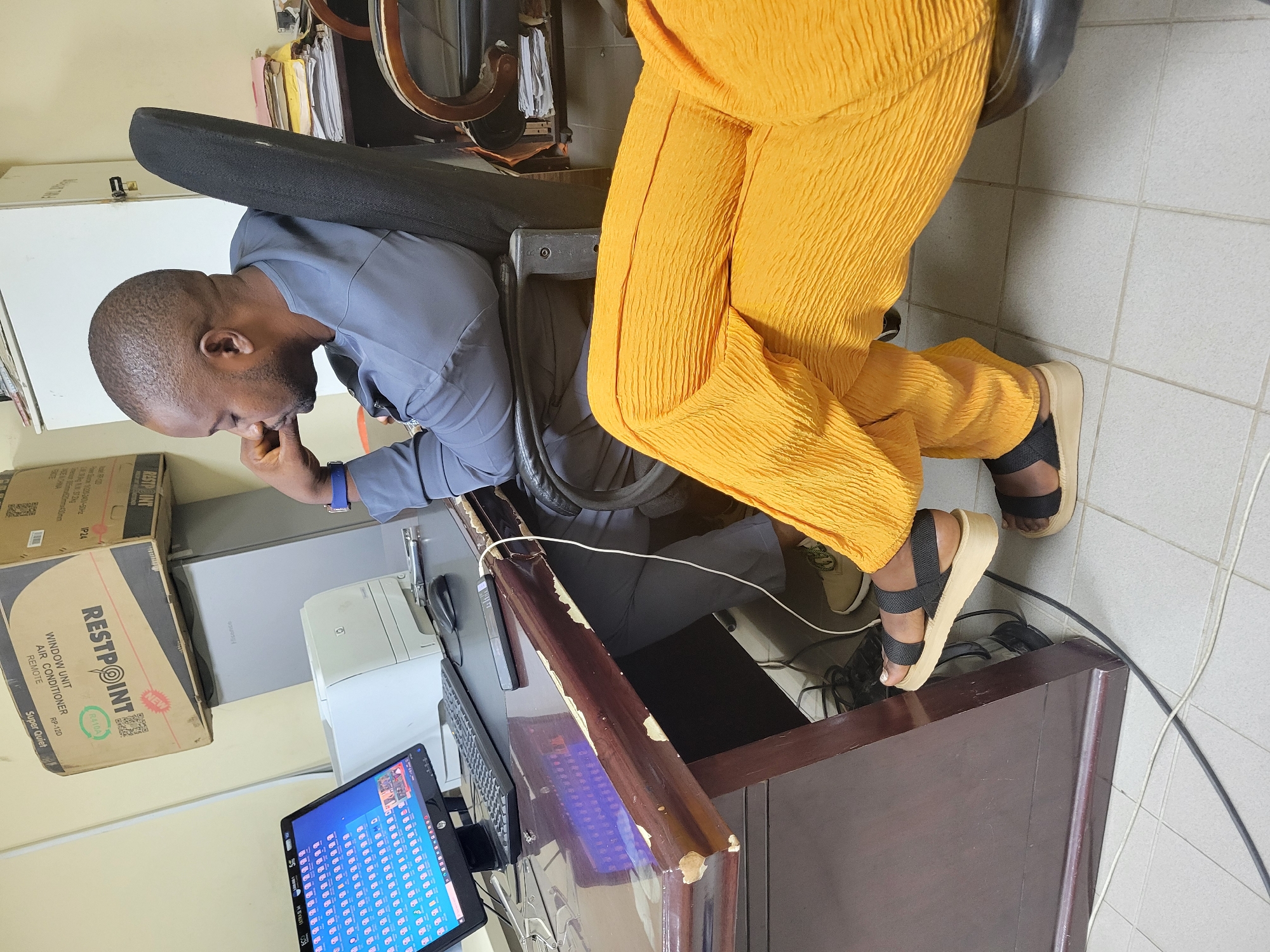 The image depicts an indoor scene, likely an office, in Abuja, Nigeria. A man in a grey shirt and pants is seated in a black office chair, facing a desk. He is resting his chin on his hand, appearing deep in thought. On the desk are a computer monitor displaying numerous file icons, a keyboard, a mouse, a printer, and a mobile phone. To the right of the man, another person is visible, wearing bright orange, textured pants and black platform sandals. The desk itself is made of dark wood and shows signs of wear and tear. In the background, filing cabinets and stacks of papers are present, contributing to the cluttered office environment. A large cardboard box for a "RESTPOINT WINDOW UNIT AIR CONDITIONER" is stacked on top of a refrigerator or a similar appliance in the upper left portion of the image. The floor is tiled.