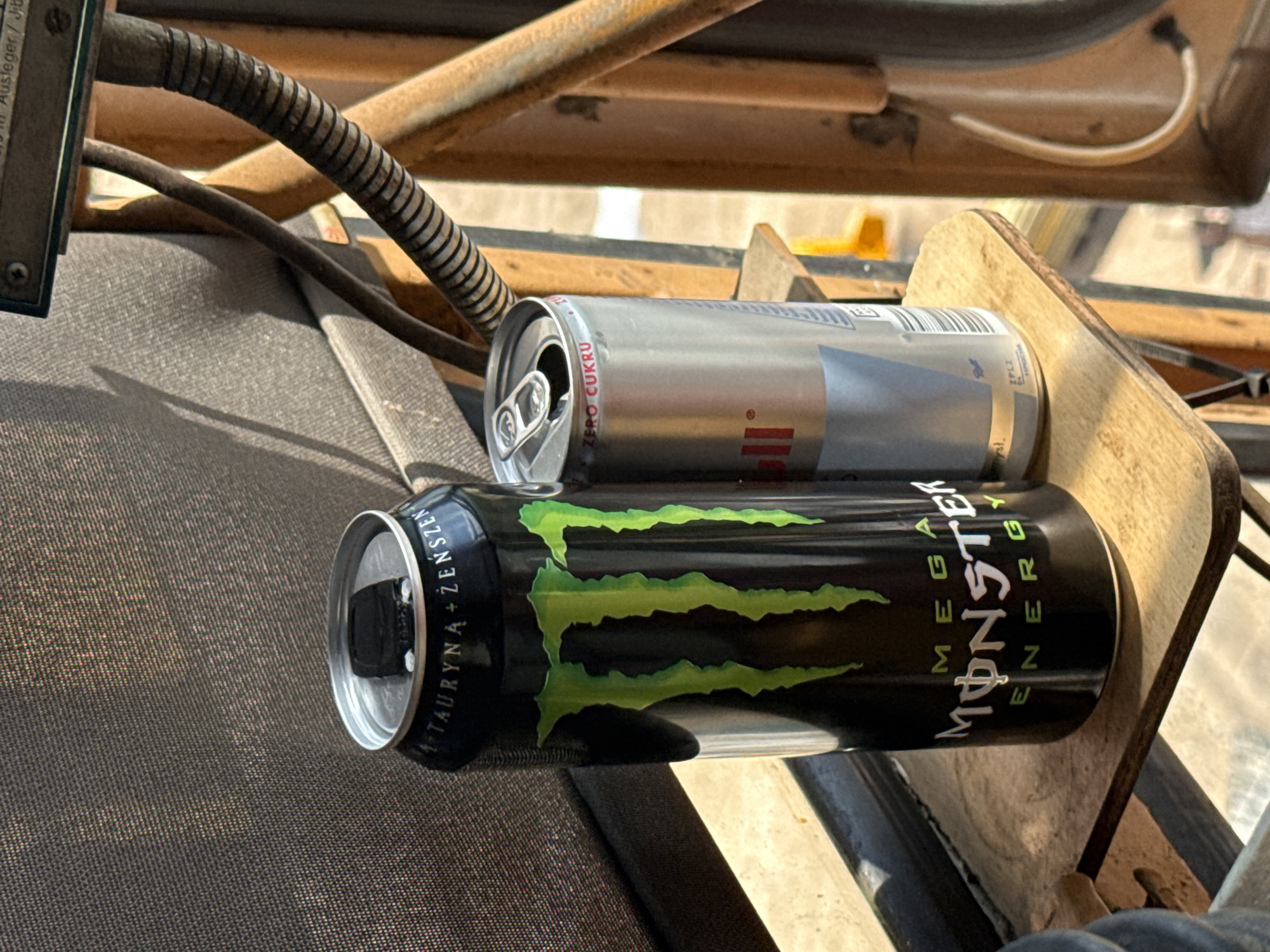 The image shows two cans of energy drinks placed on a textured, dark surface, possibly a seat or fabric covering. The can in the foreground is a black Monster Energy can with the distinctive green claw mark logo and white text reading "MEGA MONSTER ENERGY". The text on the side of the can includes "TAURYNA + ŻEŃ-SZEŃ" in white lettering. The can in the background is a silver can, possibly a Red Bull, with a white tab. Text on this can includes "ZERO CUKRU". Both cans appear to be open. The cans are resting on a wooden surface. In the background, there are metal and wooden structures, suggesting an outdoor or industrial setting. A corrugated tube and a thin wire are visible, adding to the sense of a functional or mechanical environment. The lighting suggests it is daytime, with sunlight casting shadows. There are no people visible in the image. The scene appears to be in Mikołajki, Poland.