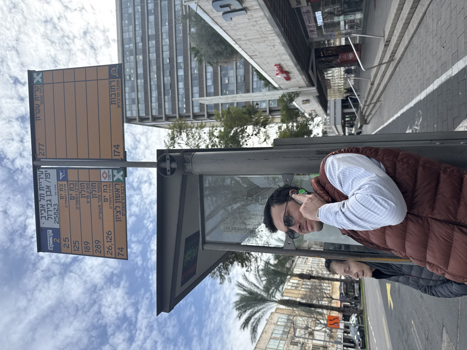 The image depicts an urban street scene at a bus stop in Tel-Aviv, Israel, specifically identified by signage as "Jaffa / Ibn Gabirol." In the foreground, a man, appearing to be in his 30s or 40s, with dark hair, dark-rimmed glasses, a light blue striped shirt, and a reddish-brown puffer vest, stands leaning against a metal bus stop pole. He is holding a smartphone to his right ear with his right hand, engaged in a phone call, with a small green earpiece visible near his ear. His gaze is directed slightly upwards and to the right. Partially obscured behind and to the left of the man is an older woman with dark, pulled-back hair, wearing a dark puffer jacket, facing to the left and seemingly waiting at the bus stop.
Above them, a blue and orange bus stop sign is mounted on the pole, displaying Hebrew text including "Tel Aviv Municipality" and bus route numbers (e.g., 2, 25, 125, 189, 289, 26, 126, 74, 277, 174) with associated destinations such as Bat Yam, Holon, Rishon LeZion, Kiryat Ekron, and Rehovot. The bus stop shelter itself features a dark grey frame and transparent panels, with a partially visible electronic display showing green text.
To the left, across a multi-lane street, several low-rise buildings and multiple palm trees are visible. Vehicles, including a bus, are present on the street. To the right of the bus stop, a commercial building with a textured facade, awnings, and visible storefronts (including "Samsung") is present, with a distinct vertical white architectural element. Beyond this, a taller, multi-story building with numerous square windows rises against the sky. The sky is partly cloudy with patches of blue. The sidewalk in the immediate foreground is paved with grey pavers, and a bicycle lane marking is faintly visible.